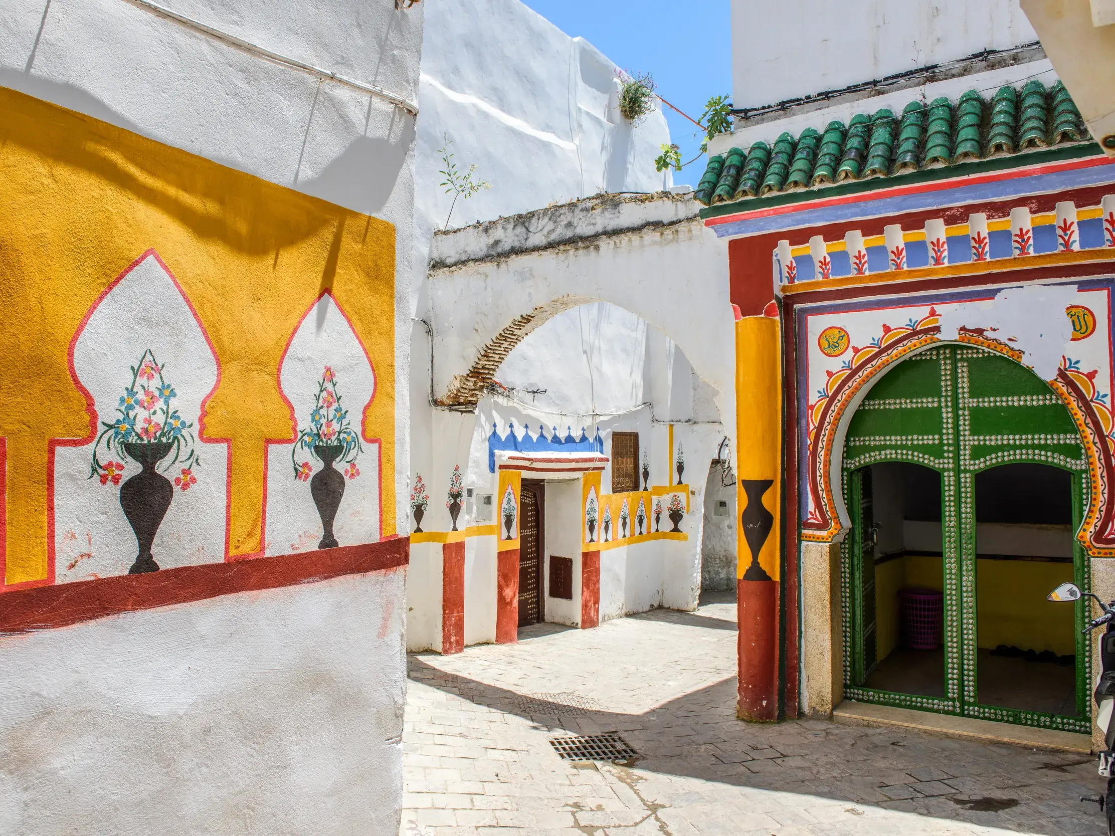 Sonnige Gasse in der Medina von Tétouan mit weiß getünchten Häusern, bunten maurischen Ornamenten und einer grünen Tür.