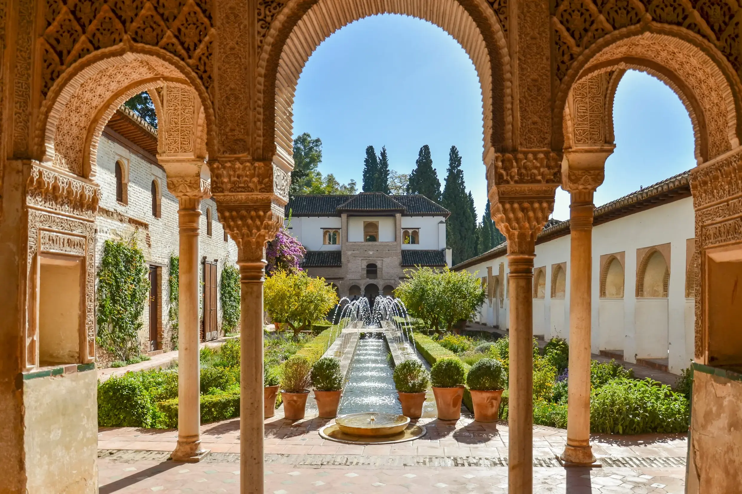 Blick durch die filigranen Bögen der Nasridenpaläste in einen sonnigen Innenhof mit Garten und Wasserspielen in der Alhambra.