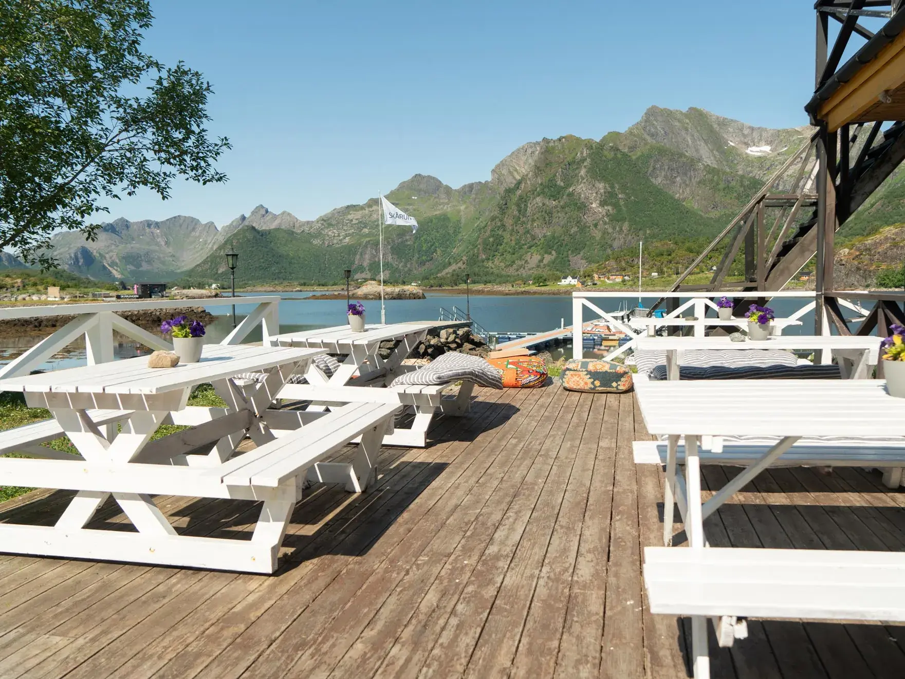 Holzterrasse mit weißen Picknicktischen am Fjord vor der beeindruckenden Bergkulisse der Lofoten in Norwegen.