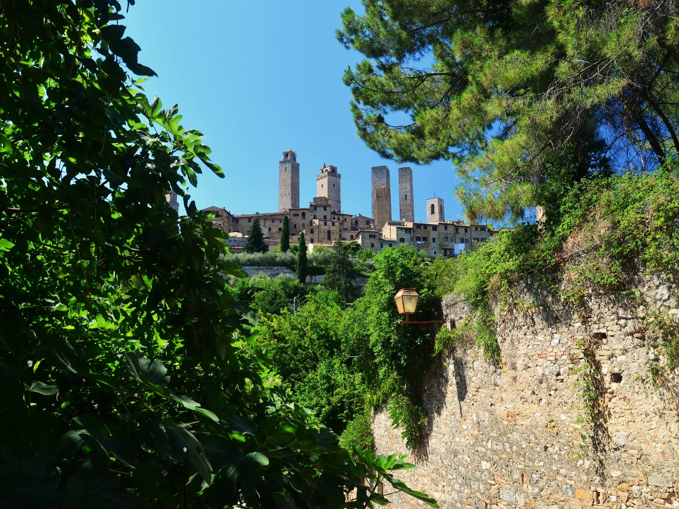 Blick von einem Wanderpfad der Via Francigena durch grüne Bäume auf die mittelalterlichen Türme von San Gimignano in der Toskana.