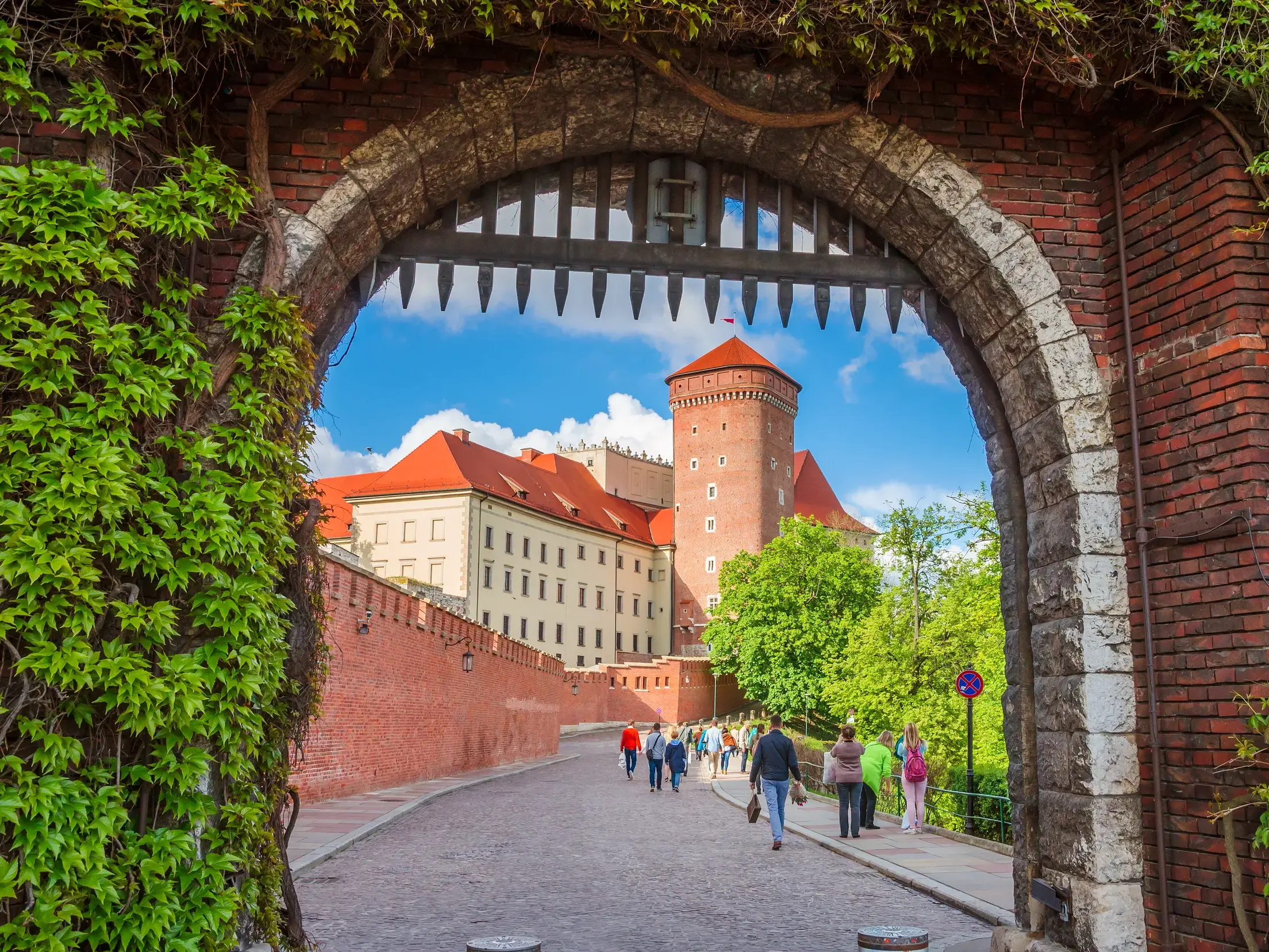Efeubewachsener Torbogen mit Blick auf den gepflasterten Weg zum Schlossberg Wawel in Krakau, auf dem Besucher spazieren.