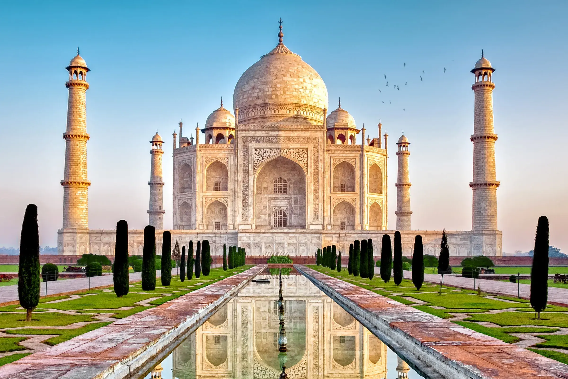 Märchenhaftes Taj Mahal in Agra bei Sonnenaufgang, dessen weiße Fassade sich im langen Wasserbecken der Gärten spiegelt.