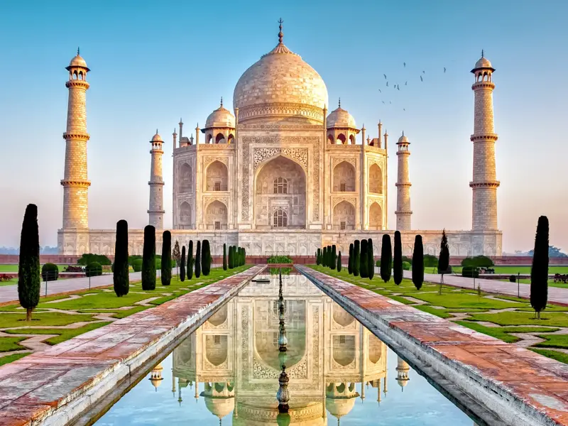 Märchenhaftes Taj Mahal in Agra bei Sonnenaufgang, dessen weiße Fassade sich im langen Wasserbecken der Gärten spiegelt.