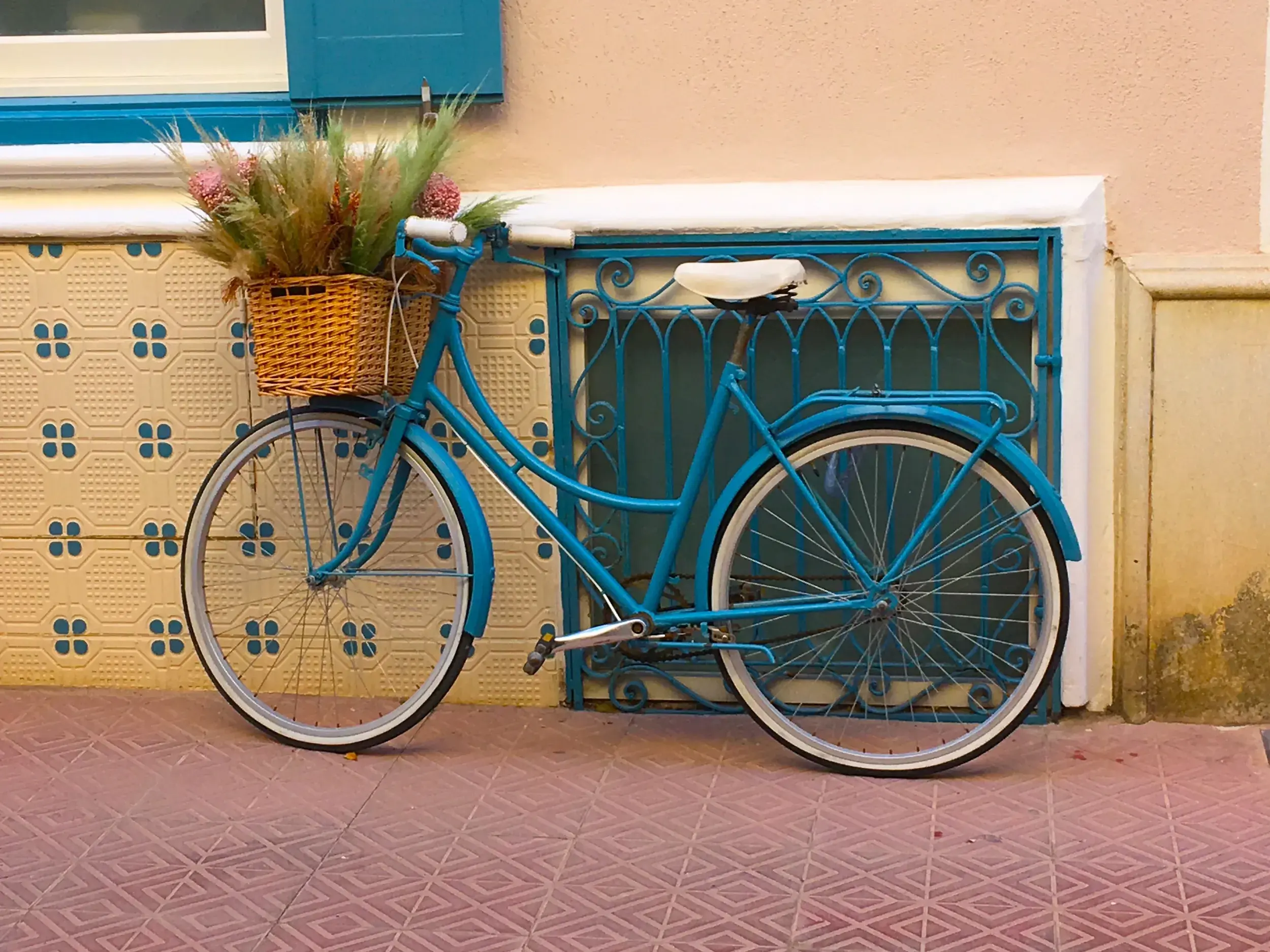 Hellblaues Retro-Fahrrad mit Blumenkorb vor einer farbenfrohen Hausfassade in den Gassen von Ciutadella auf Menorca.