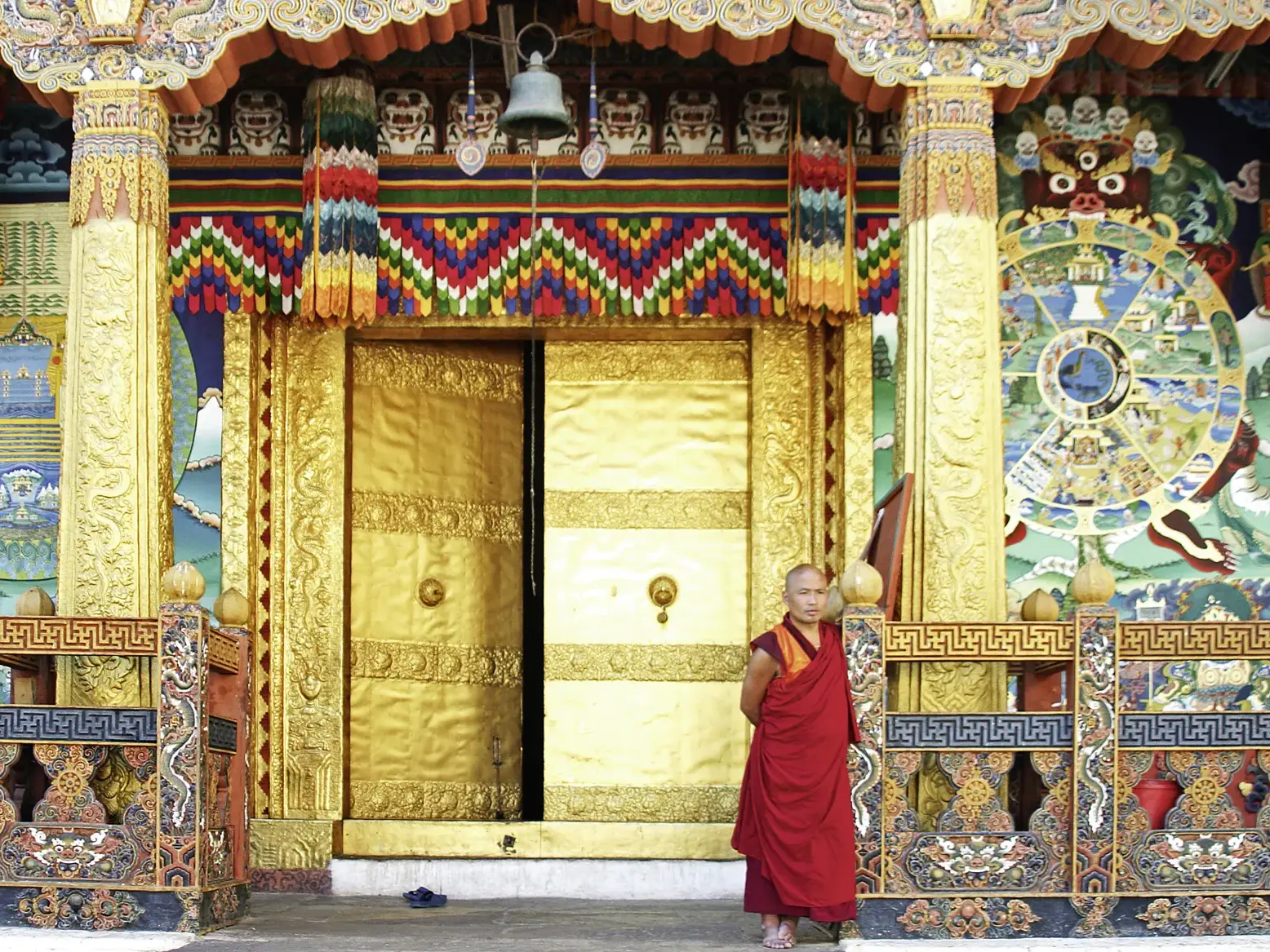 Junger Mönch in roter Robe steht vor dem reich verzierten, goldenen Tor eines buddhistischen Klosters in Bhutan.