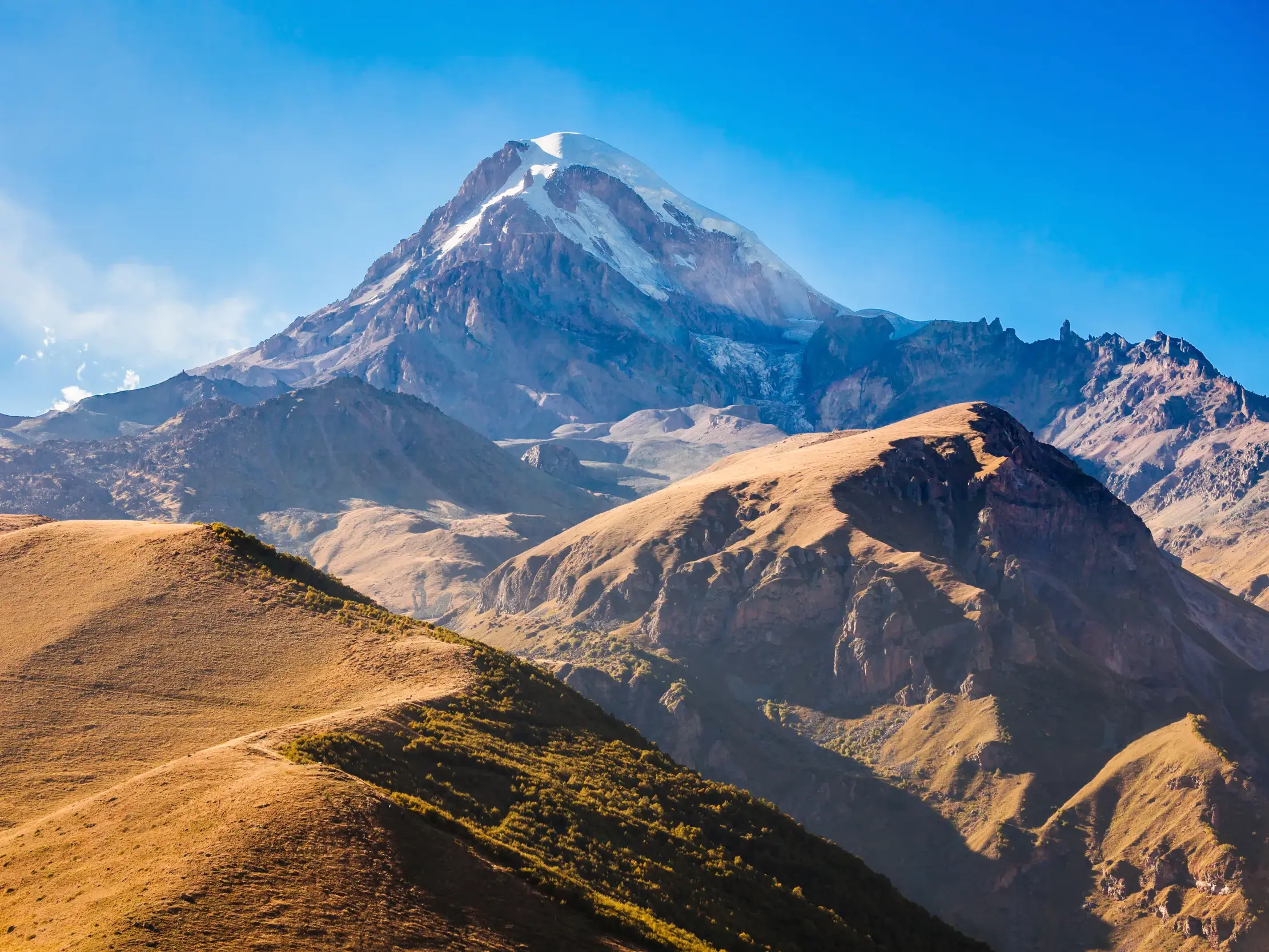 Wanderweg über sonnige, goldene Hügel mit Blick auf den majestätischen, schneebedeckten Berg Kasbek in Georgien.