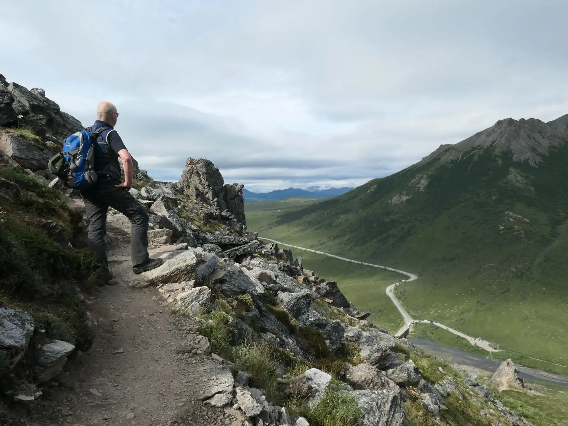 Wanderer auf dem felsigen McLaren Summit Trail blickt auf den Denali Highway, der sich durch ein weites, grünes Tal schlängelt.