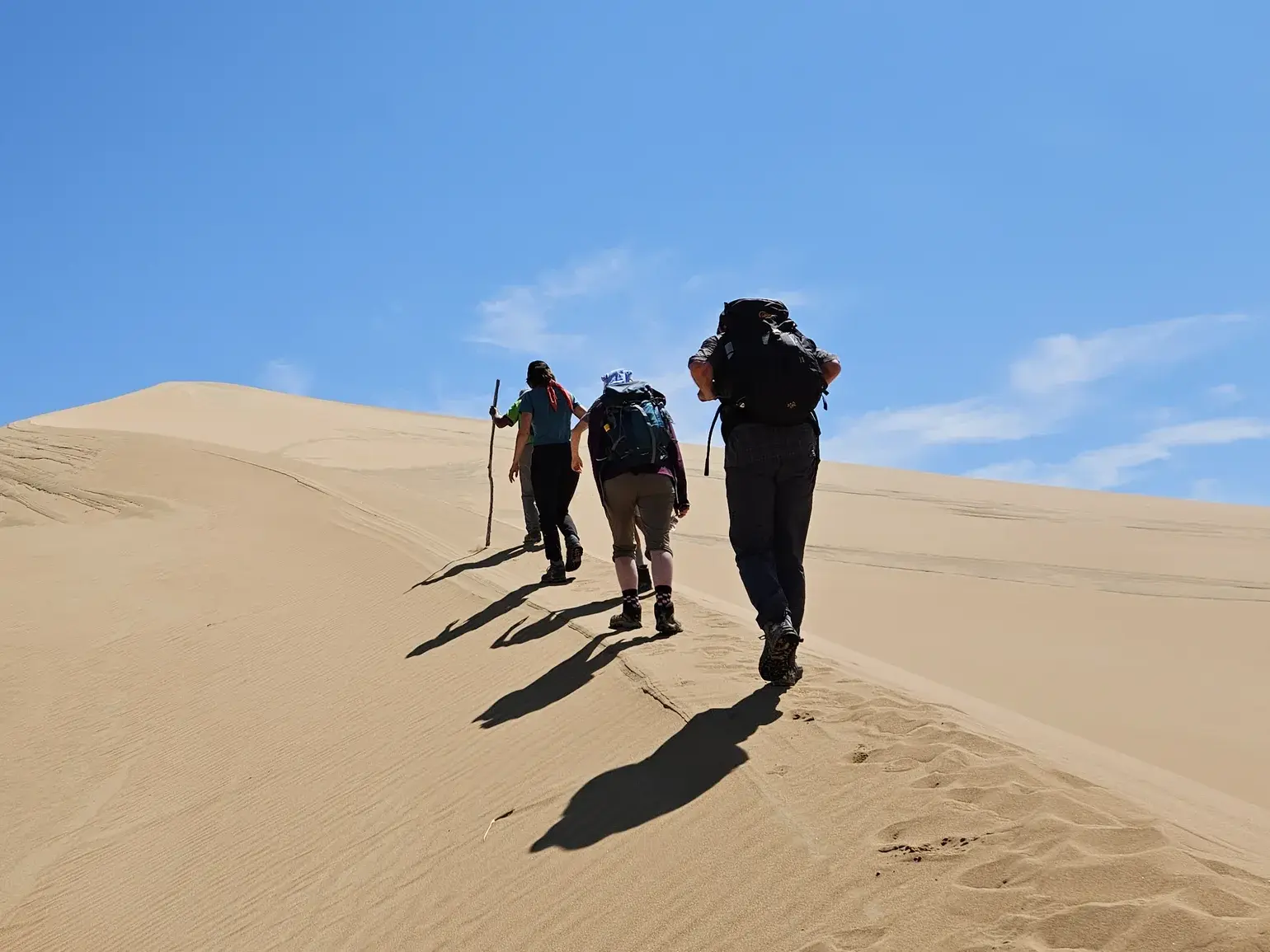 Drei Trekking-Reisende mit Rucksäcken wandern über den sonnigen Grat einer Sanddüne im Dünenfeld Mongol Els in der Mongolei.