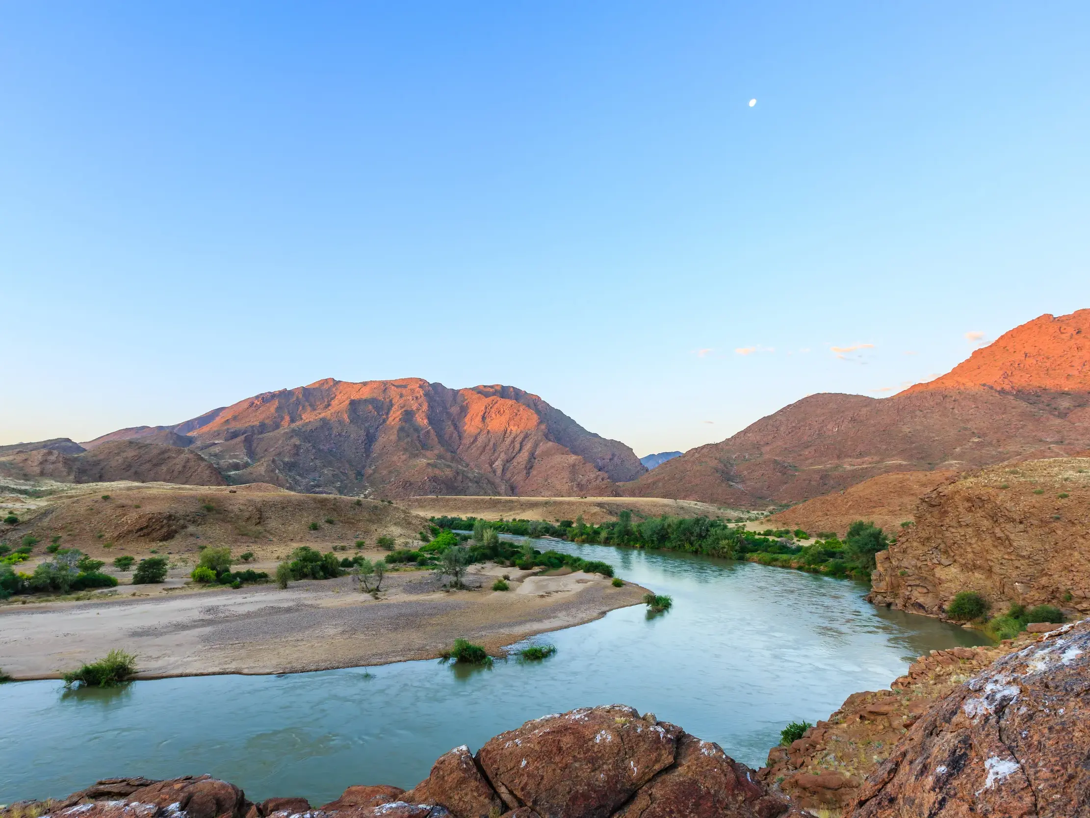 Der breite Kwanza-River schlängelt sich durch eine karge Berglandschaft in Angola, beleuchtet vom warmen Abendlicht.