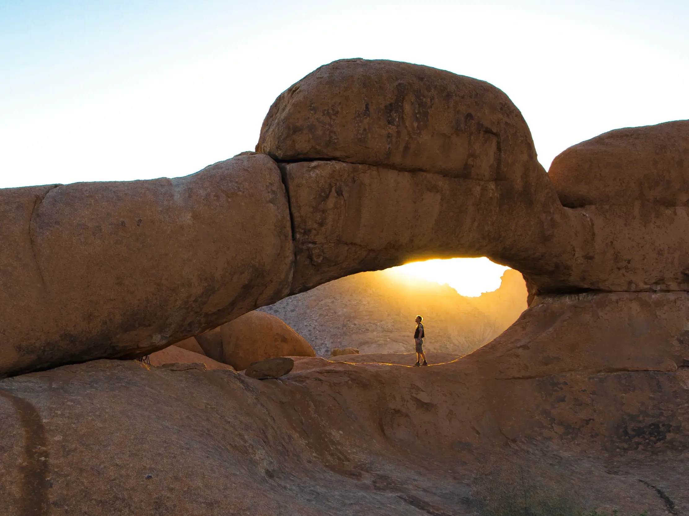 Ein Reisender steht im goldenen Licht der untergehenden Sonne unter einem markanten Felsbogen in Namibia.