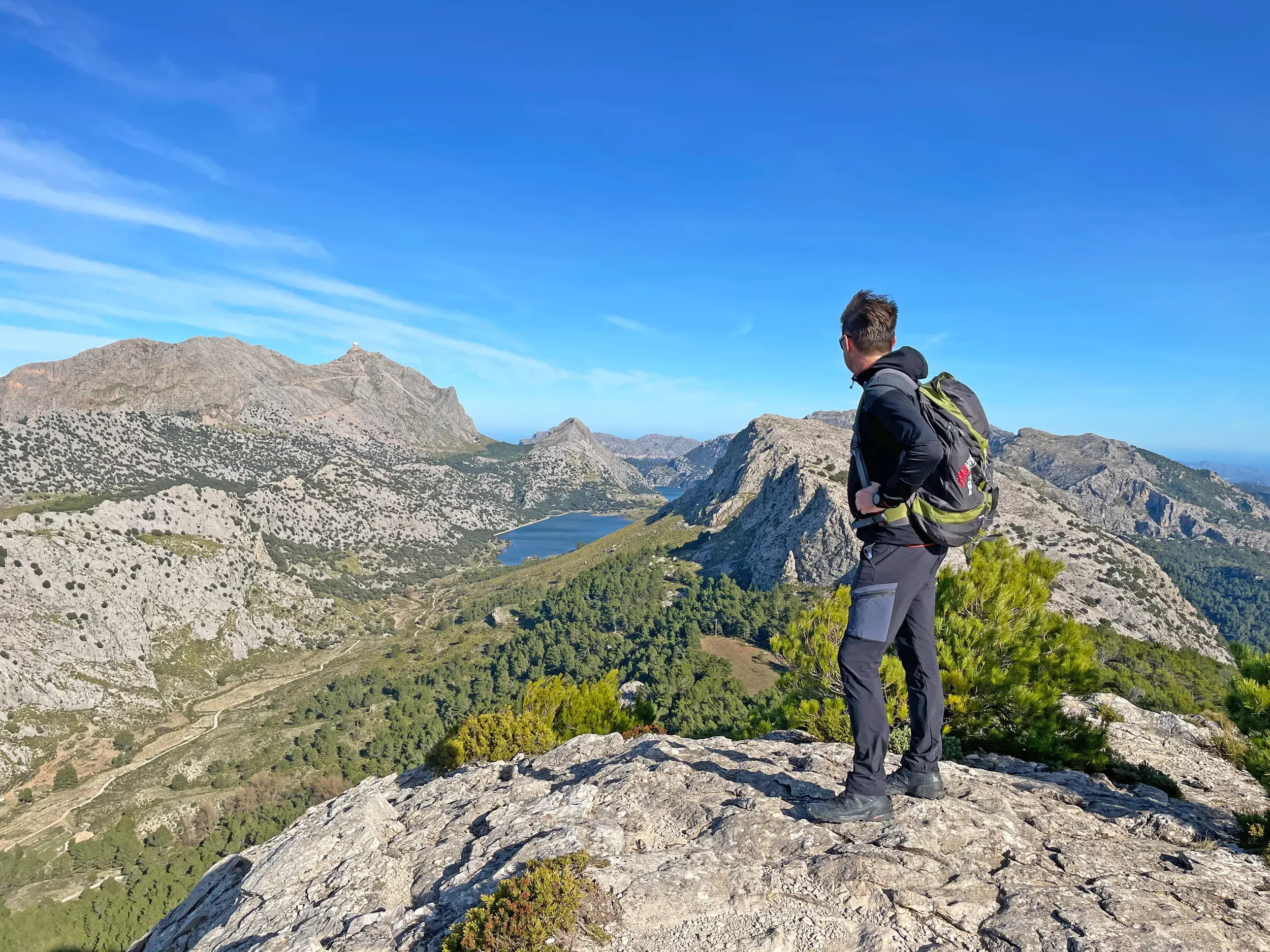 Ein Wanderer mit Rucksack genießt auf einem Felsgipfel den Panoramablick auf den Cuber Stausee in der Serra de Tramuntana.
