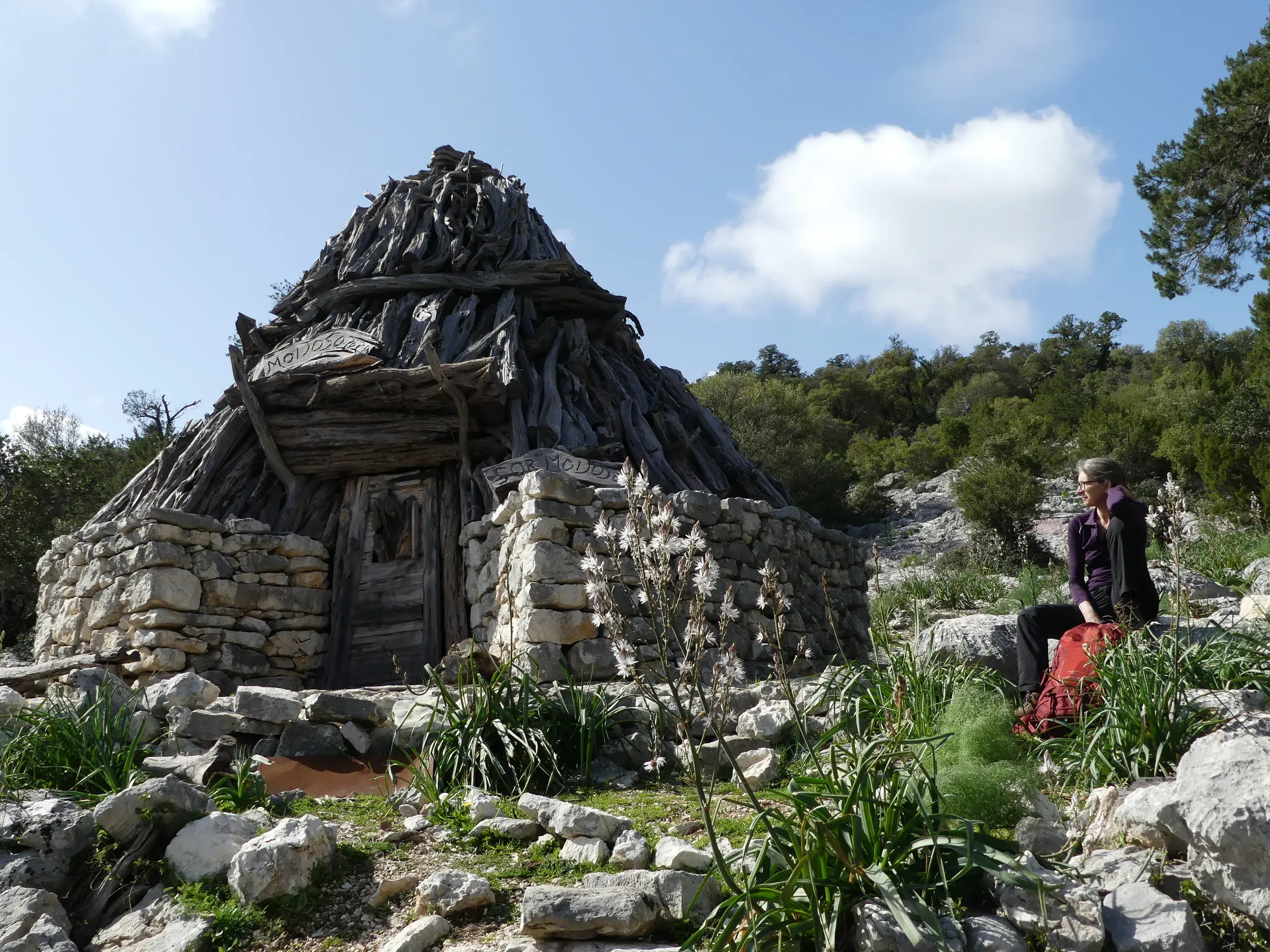 Eine Wanderin mit rotem Rucksack rast an der urigen Hirtenhütte Sos Moijos aus Stein und Holz im felsigen Supramonte Sardiniens.