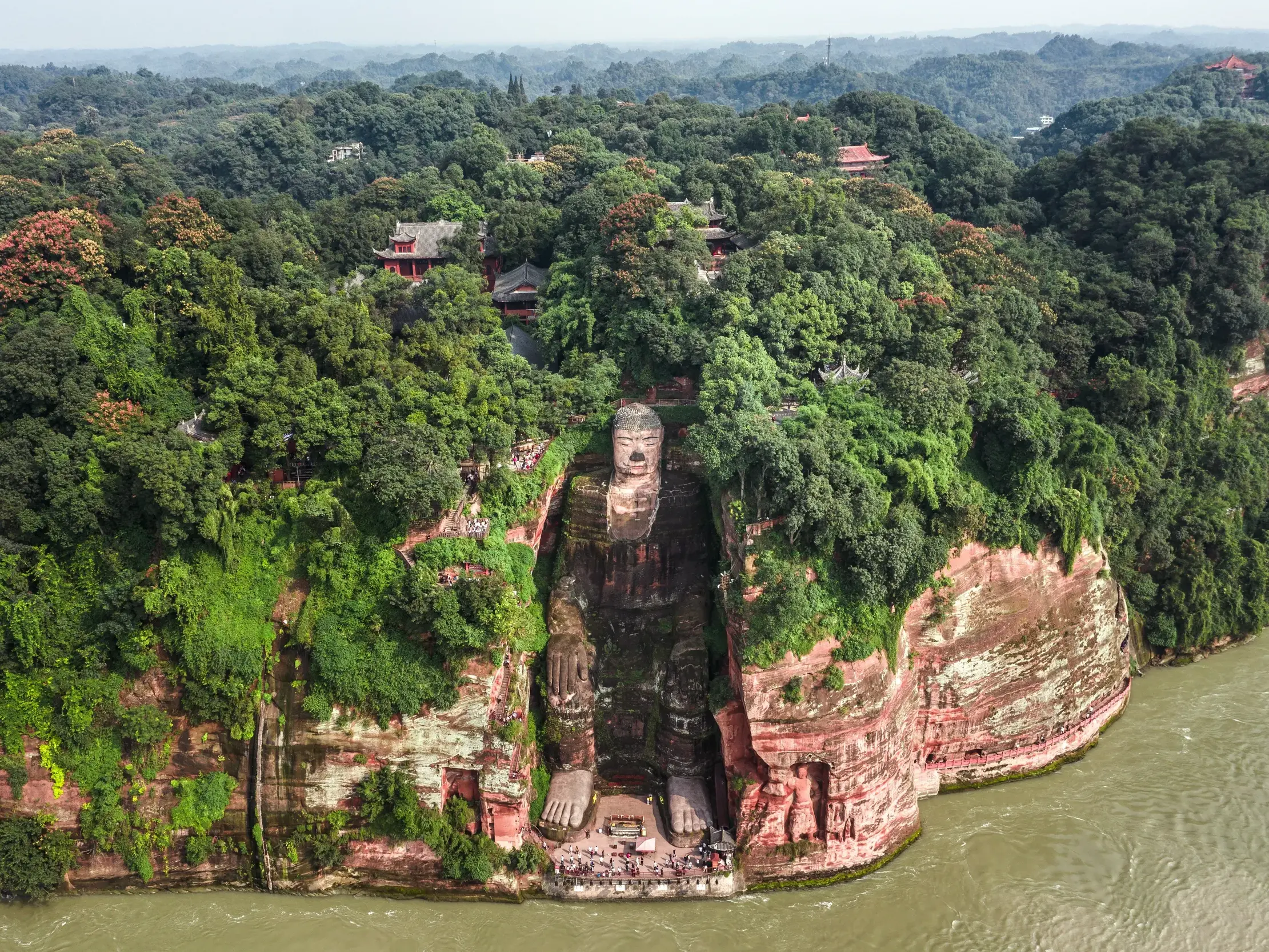 Majestätischer Großer Buddha von Leshan, in eine bewaldete Felsklippe über dem Min-Fluss gemeißelt, mit Menschen an seinen Füßen.