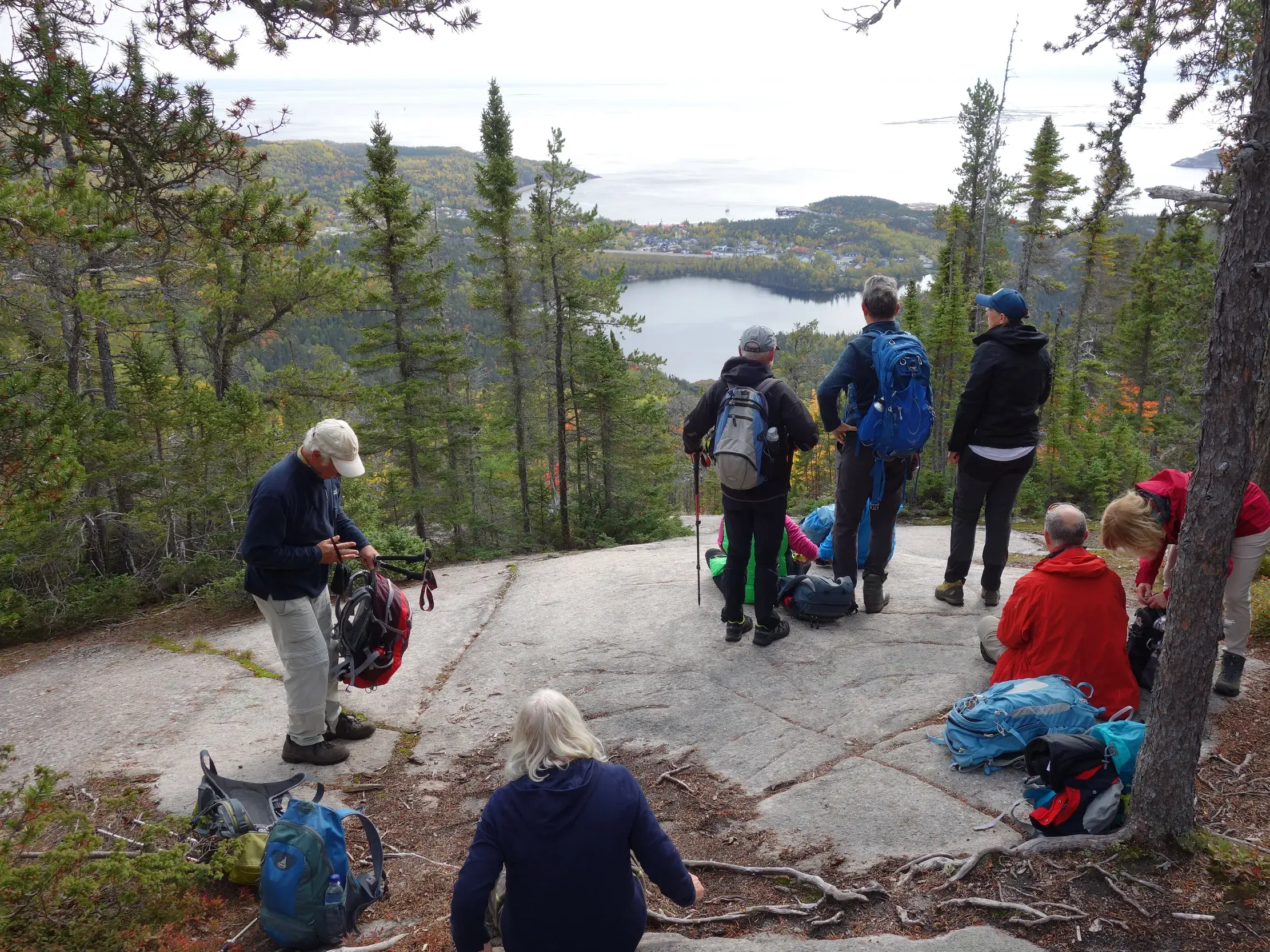 Eine Wandergruppe pausiert auf einem Felsplateau und blickt über die herbstlichen Wälder auf den Saguenay-Fjord in Québec.