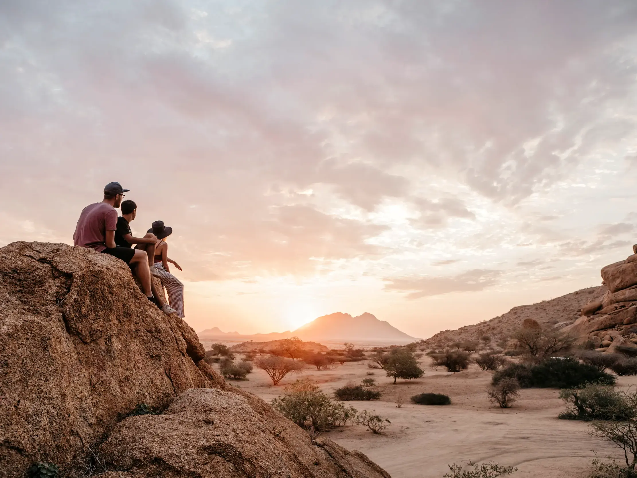 Junge Reisegruppe auf einem Felsen beobachtet den farbenfrohen Sonnenuntergang über der weiten Wüstenlandschaft Namibias.