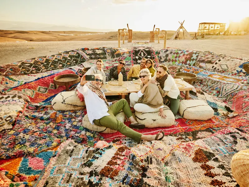 Eine fröhliche Marco Polo Reisegruppe macht ein Selfie bei einem Picknick auf bunten Teppichen in der Wüste Tunesiens.