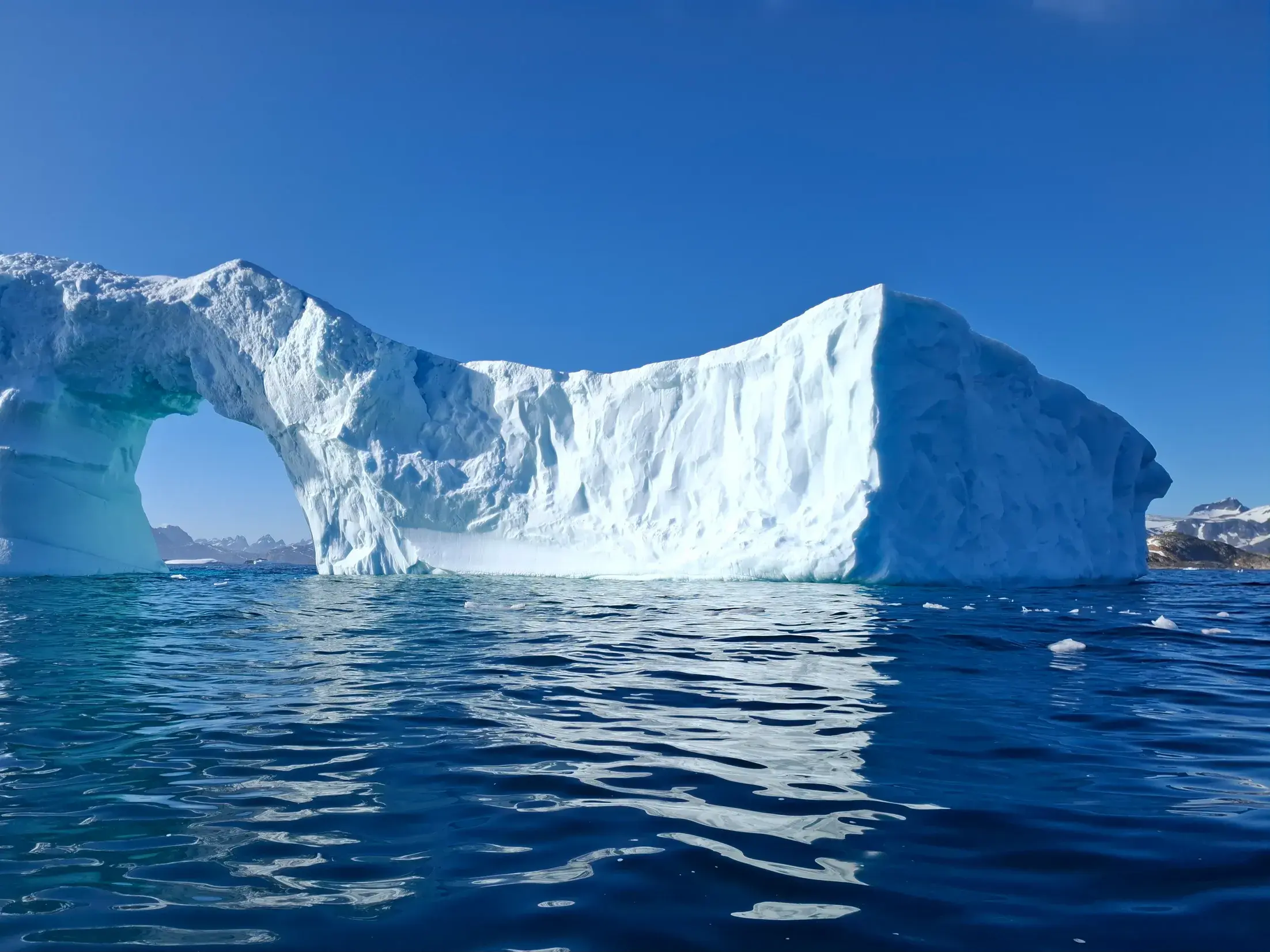 Blick von einem Boot auf einen riesigen Eisberg mit Torbogen, der im blauen Wasser des Sermilik-Fjords in Grönland treibt.