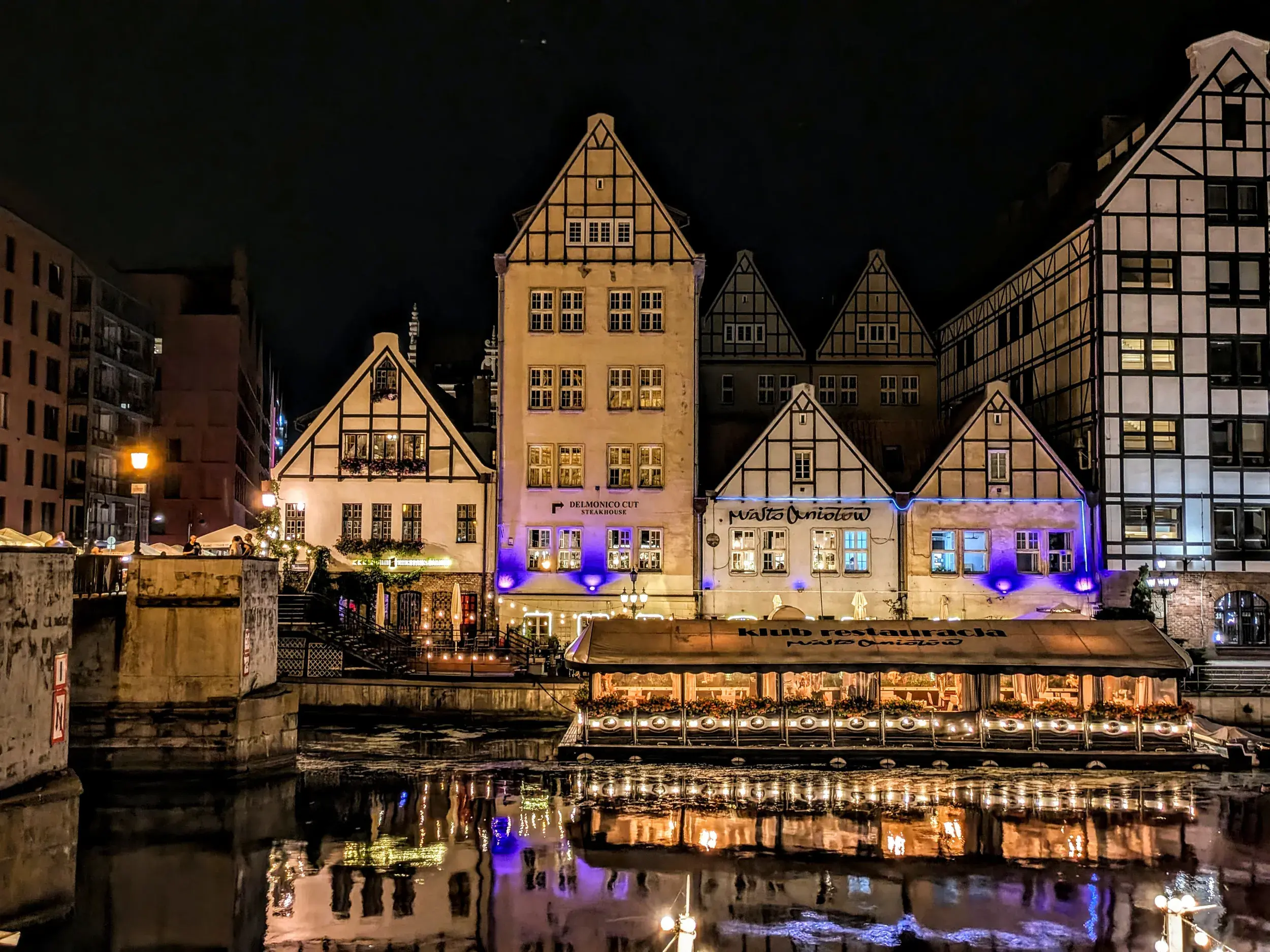 Die bunten, beleuchteten Fassaden der historischen Giebelhäuser in der Danziger Altstadt spiegeln sich nachts im Wasser der Mottlau.