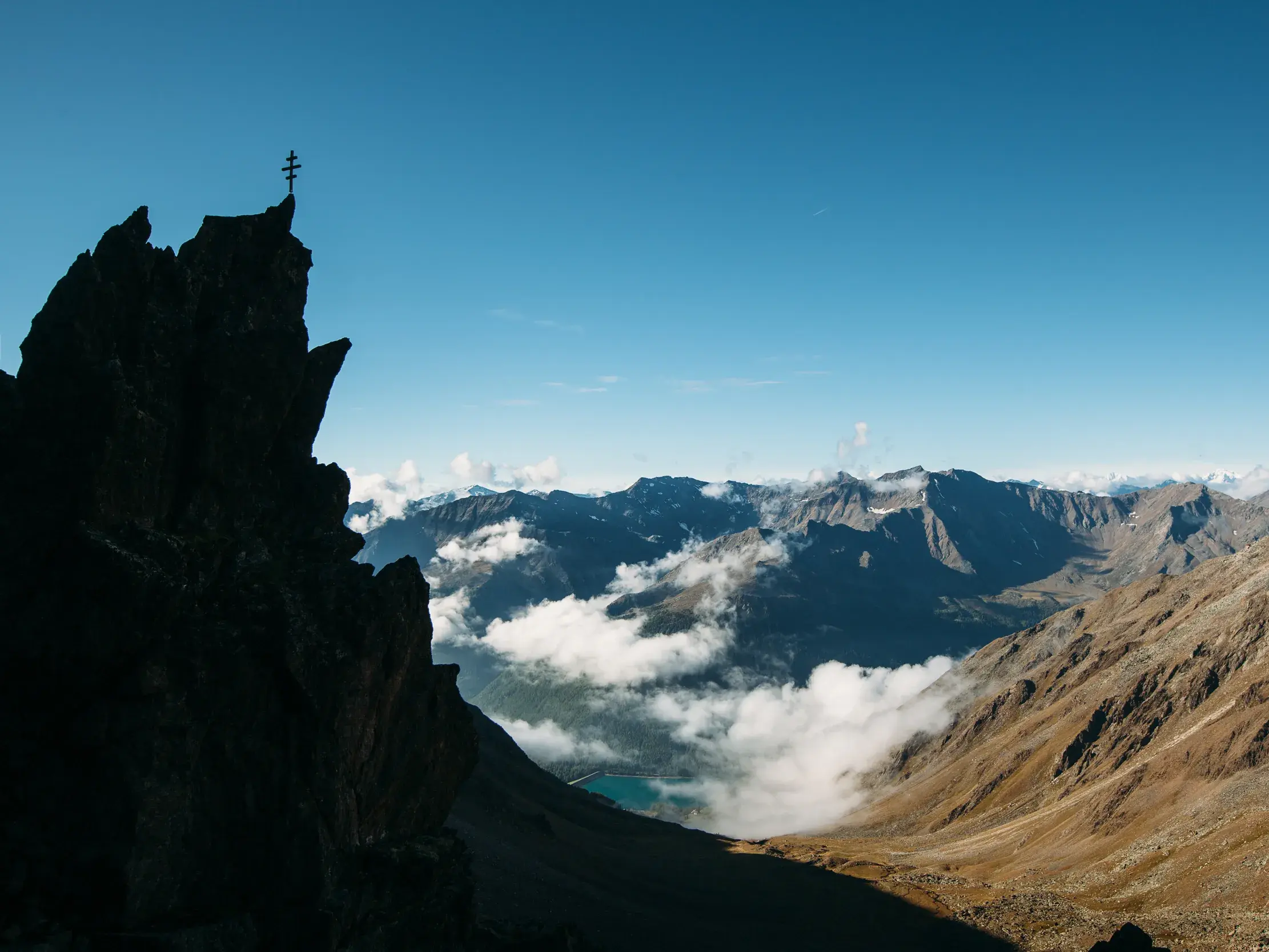 Blick von einem Gipfel mit Kreuz über ein wolkiges Tal auf den türkisblauen Vernagt-Stausee in den Ötztaler Alpen.