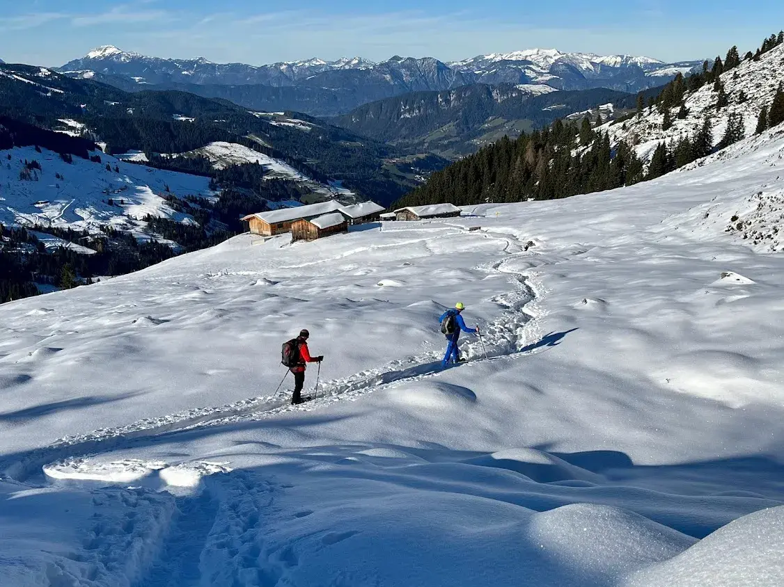 Zwei Schneeschuhwanderer auf einem tief verschneiten Hang mit Blick auf urige Almhütten und das Bergpanorama der Kitzbüheler Alpen.