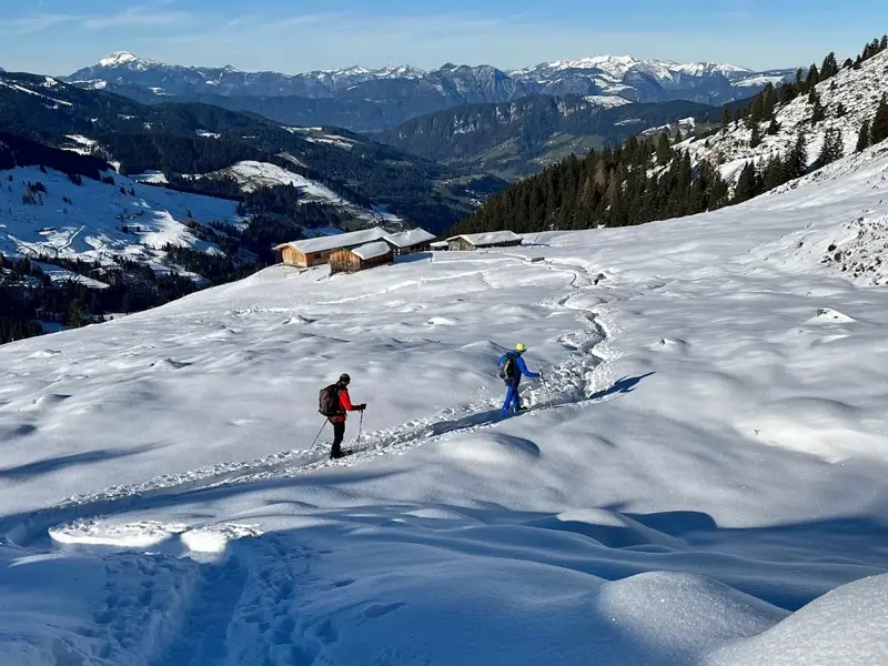 Zwei Schneeschuhwanderer auf einem Pfad im Tiefschnee vorbei an Almhütten mit Panoramablick auf die Kitzbüheler Alpen.