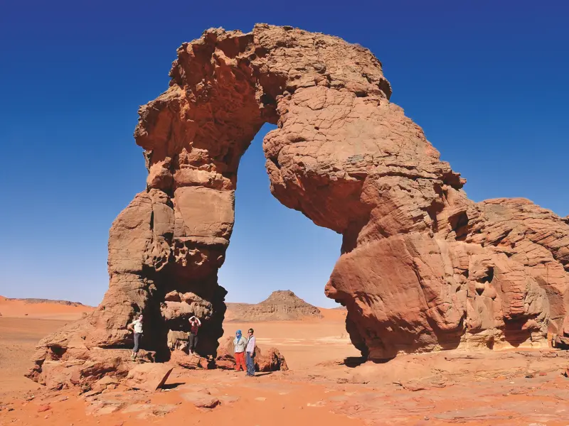 Wandergruppe pausiert vor einem imposanten Felsbogen aus rotem Sandstein während eines Trekkings im Tadrart, Algerien.