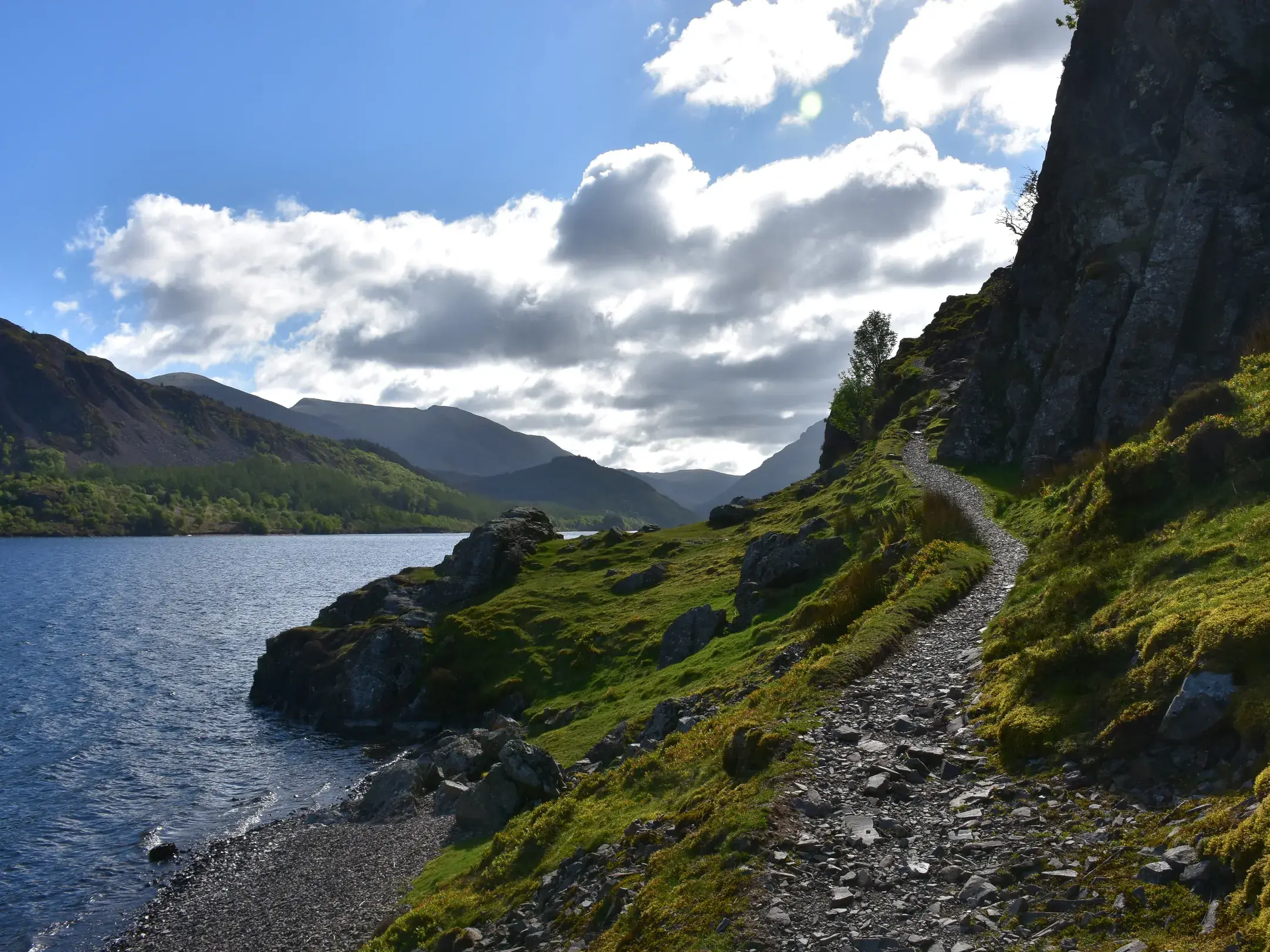 Ein steiniger Wanderpfad führt am Ufer des Ennerdale Water im Lake District entlang, im Hintergrund erheben sich Berge.