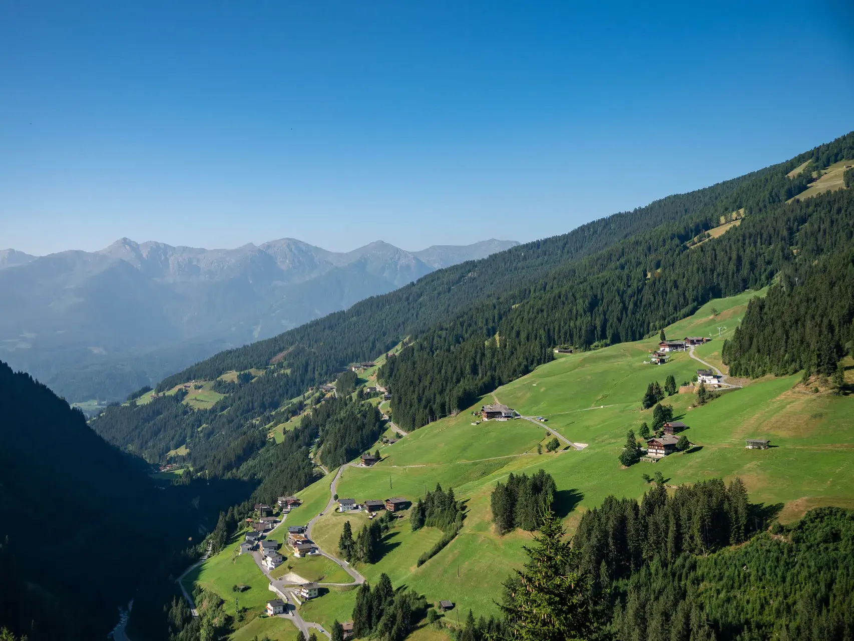Weitblick über das sonnige Villgratental mit verstreuten Bauernhöfen an grünen Hängen und den Villgrater Bergen am Horizont.