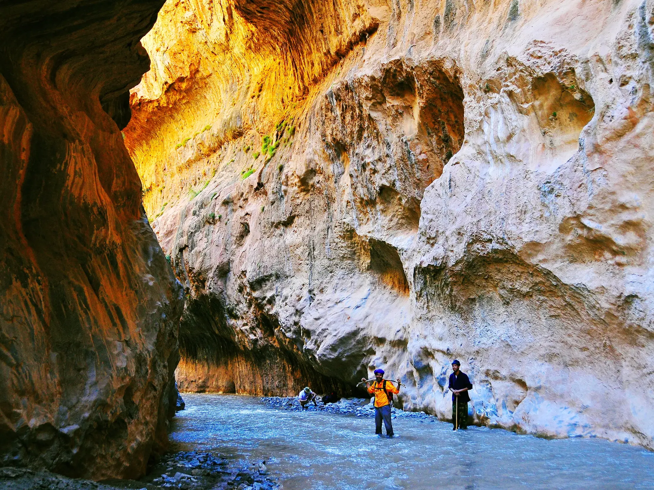 Trekkinggruppe watet durch einen Fluss in der engen Mgoun-Schlucht in Marokko, umgeben von hohen, sonnigen Felswänden.