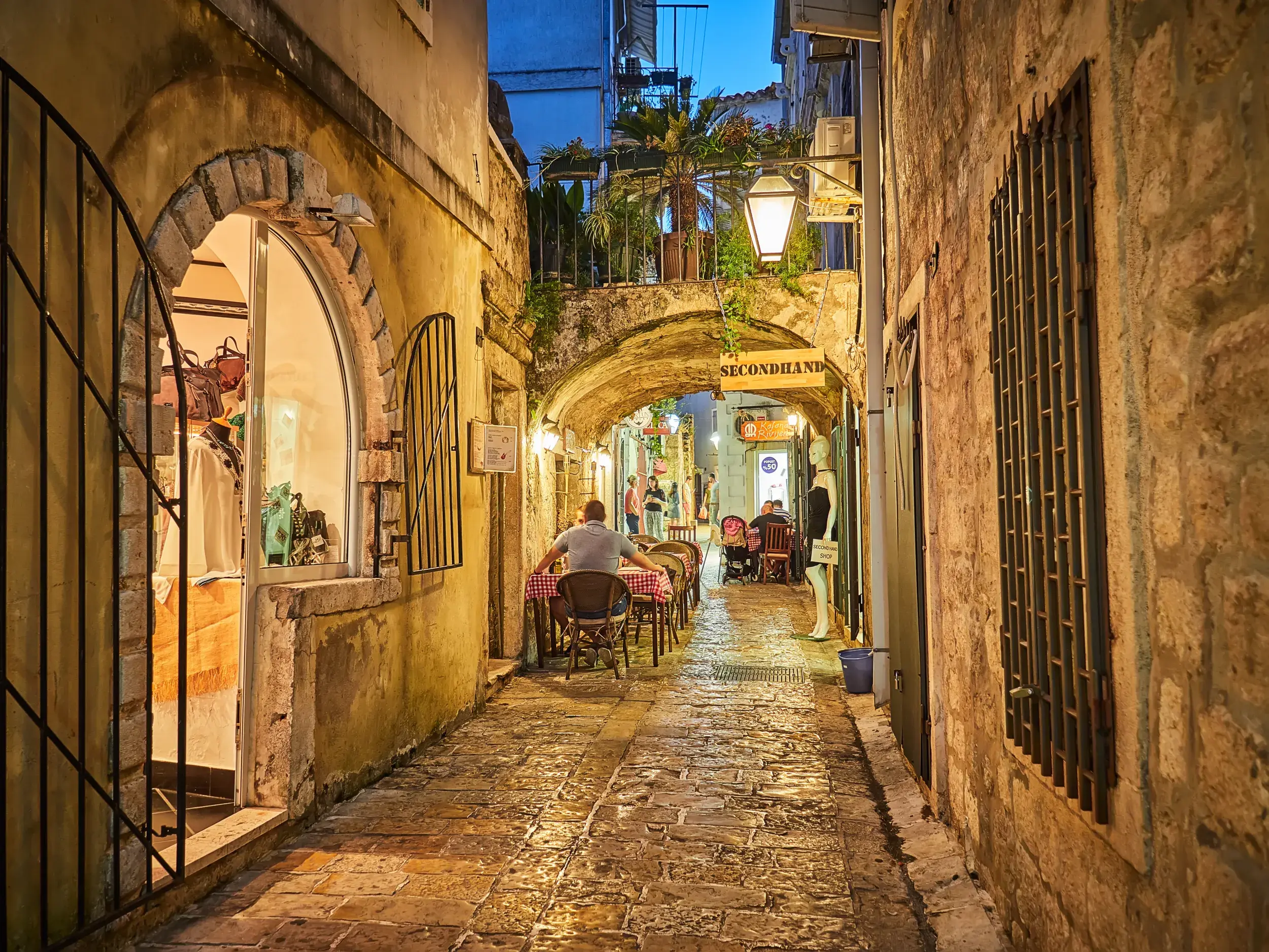 Abendstimmung in einer engen Gasse der Altstadt von Budva, wo Gäste vor kleinen Restaurants auf dem Kopfsteinpflaster sitzen.