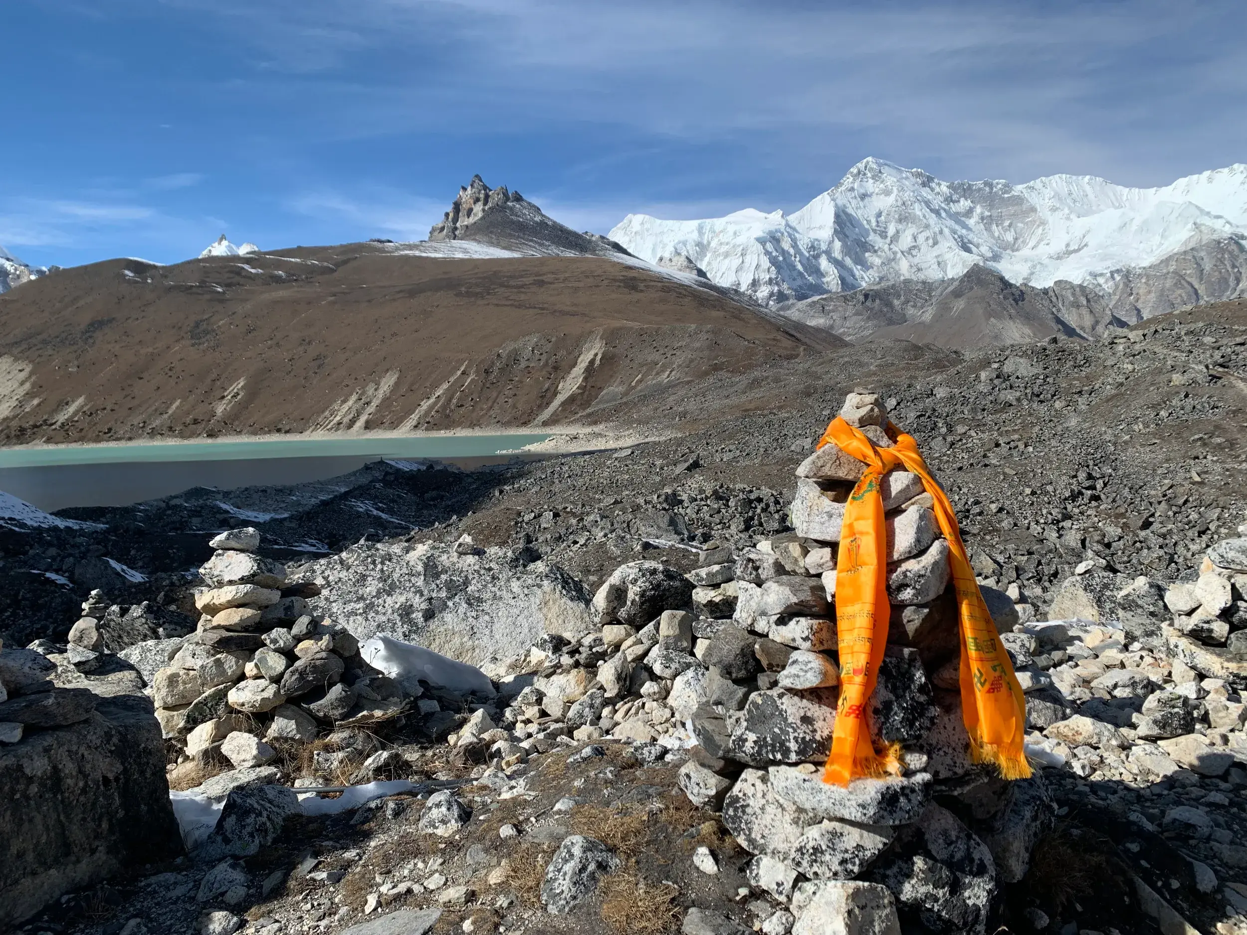 Steinmännchen mit orangem Gebetsschal am Gokyo-See in der Everest-Region vor der Kulisse schneebedeckter Achttausender.
