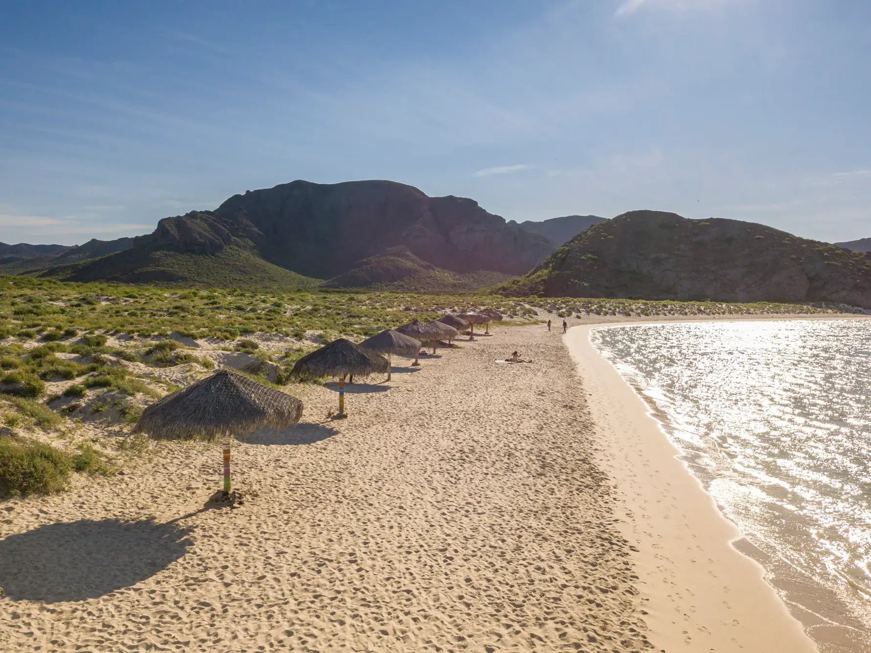 Wanderung entlang der geschützten Bucht Playa Balandra mit hellem Sandstrand, Palapa-Schirmen und der Bergkulisse von Baja California.