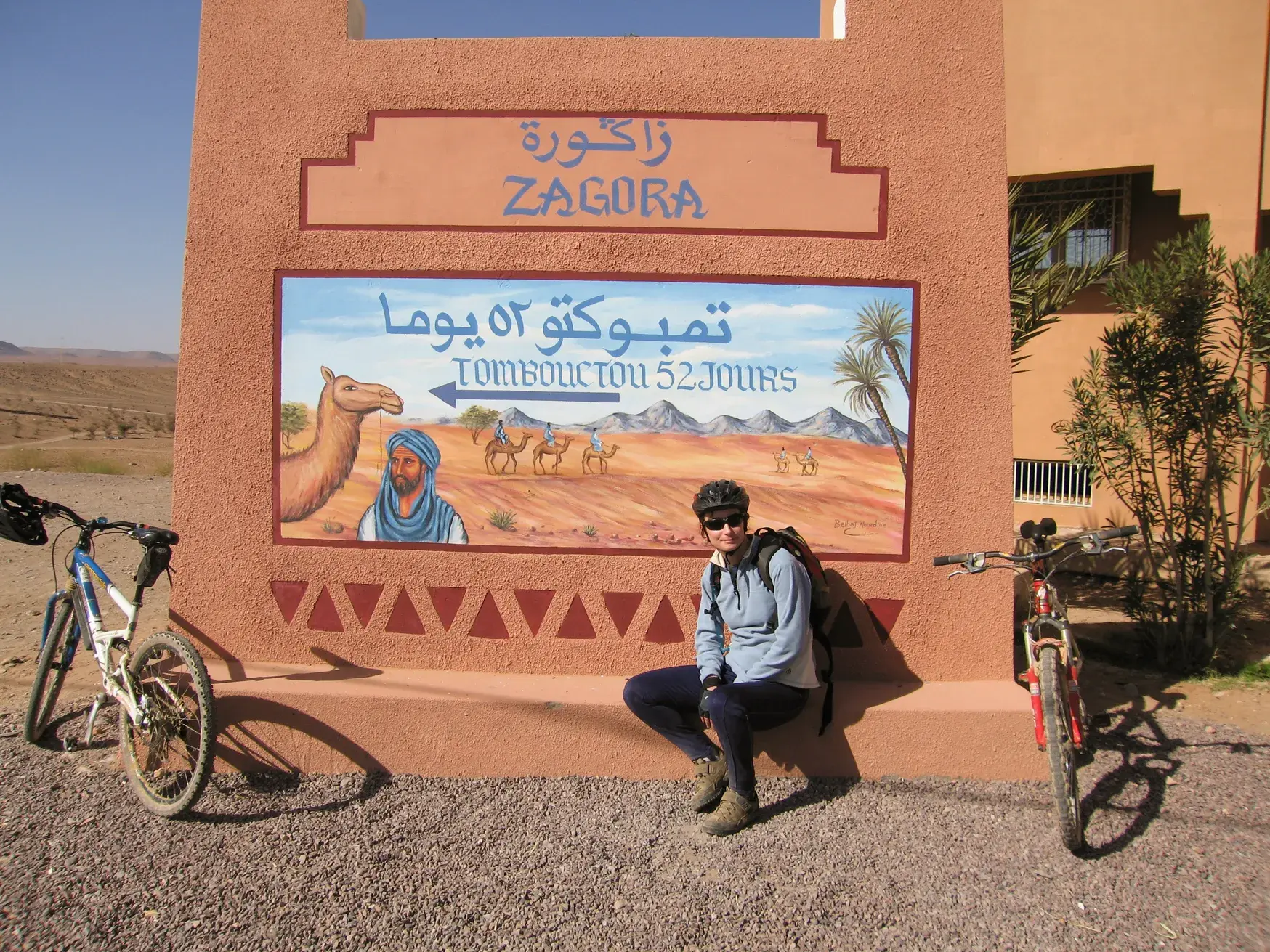 Radfahrerin mit Helm pausiert auf ihrer geführten Rundreise vor dem berühmten „Tombouctou 52 jours“-Schild in Zagora, Marokko.