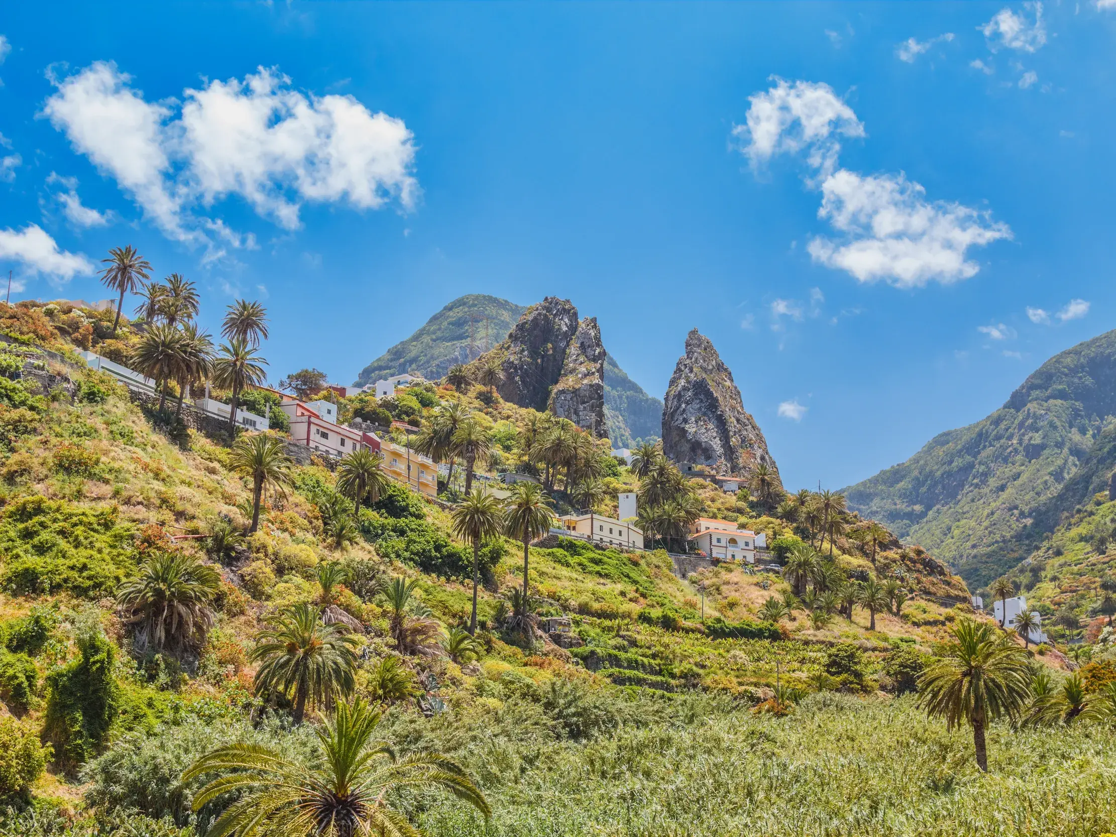 Sonniger Blick in das grüne Tal von Benchijigua auf La Gomera mit Palmen, Häusern am Hang und markanten Vulkanfelsen.