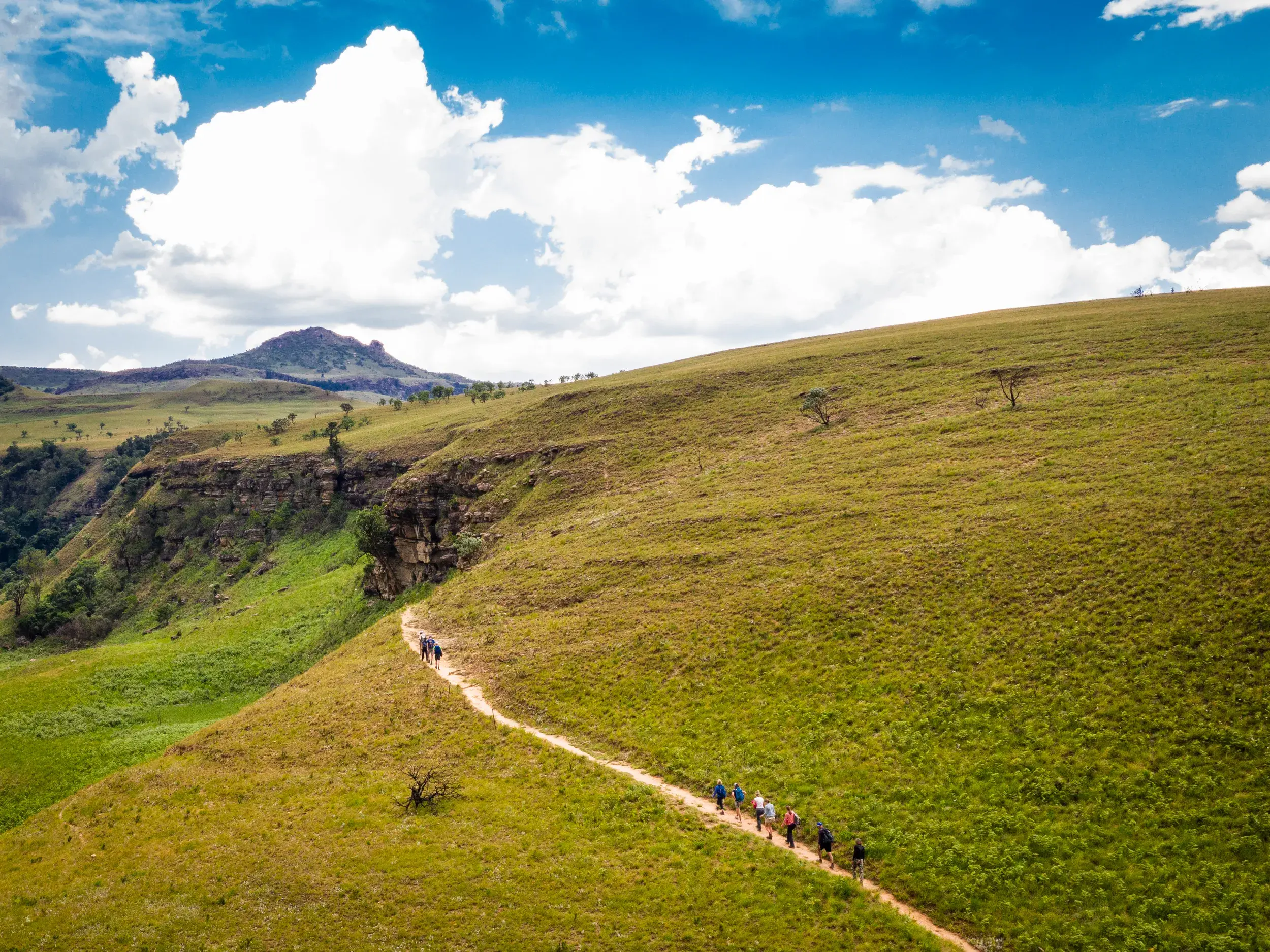 Eine Hauser-Reisegruppe wandert auf einem schmalen Pfad durch die weite, grüne Berglandschaft der Drakensberge in Südafrika.
