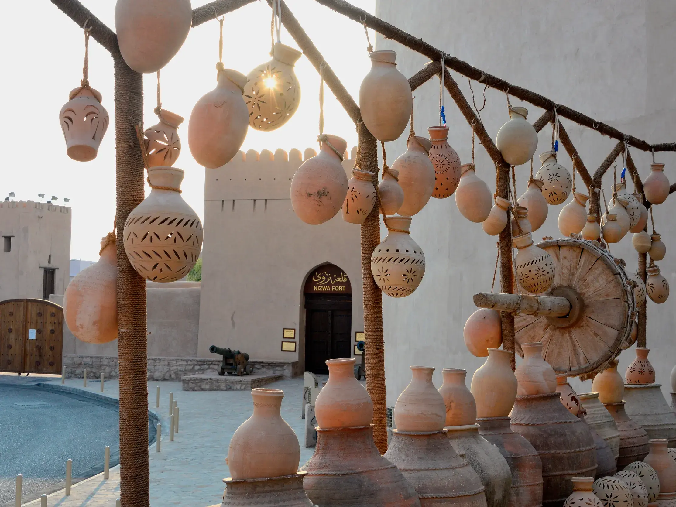 Handgefertigte Tonkrüge und Laternen hängen an einem Stand vor den Mauern des sonnenbeschienenen Nizwa Fort im Oman.