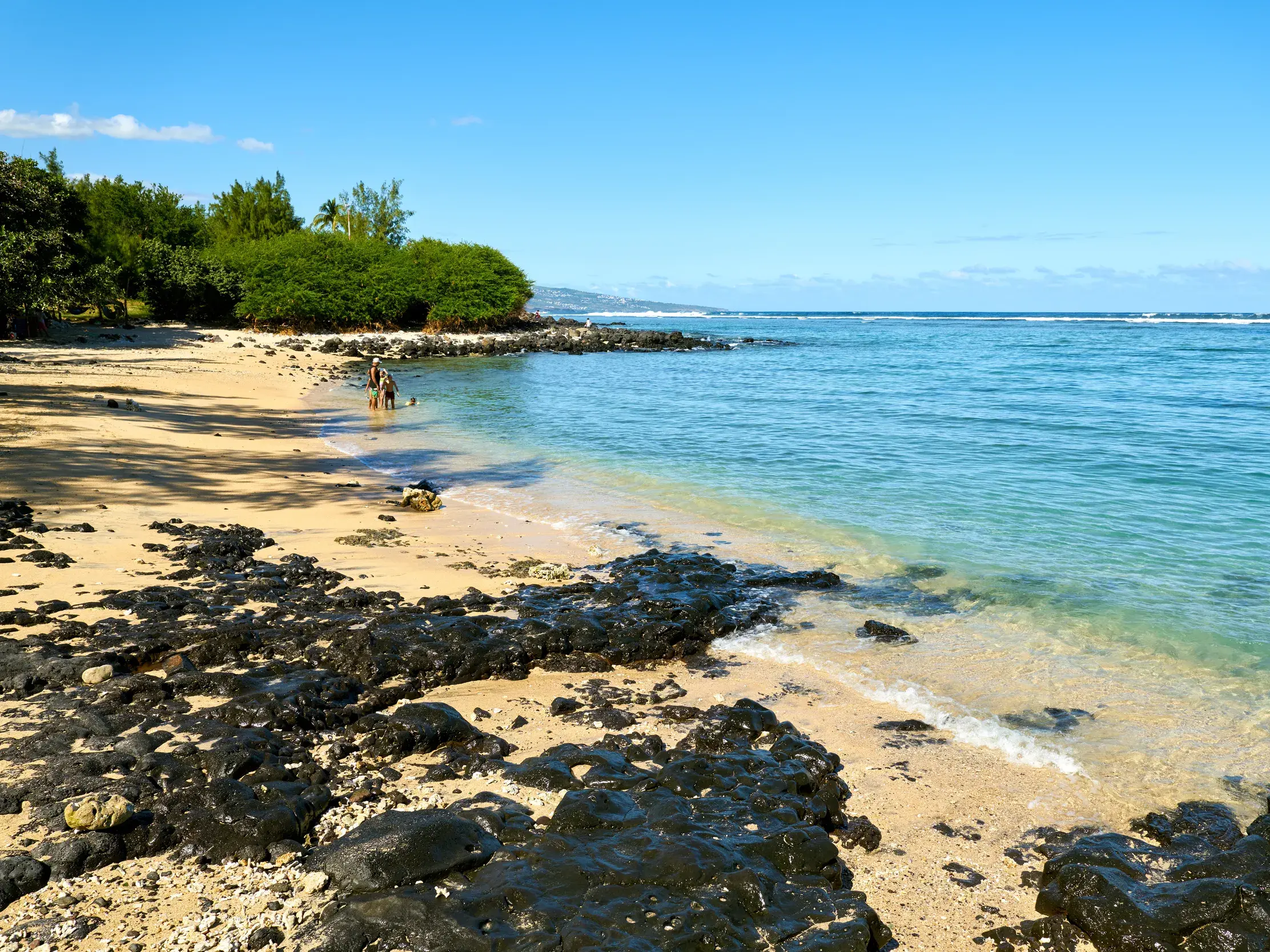 Sonniger Sandstrand mit schwarzen Vulkanfelsen auf La Réunion, wo zwei Personen im türkisen Wasser des Indischen Ozeans baden.