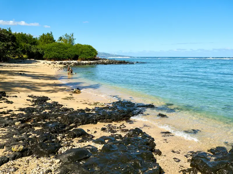 Sonniger Sandstrand mit schwarzen Vulkanfelsen auf La Réunion, wo zwei Personen im türkisen Wasser des Indischen Ozeans baden.