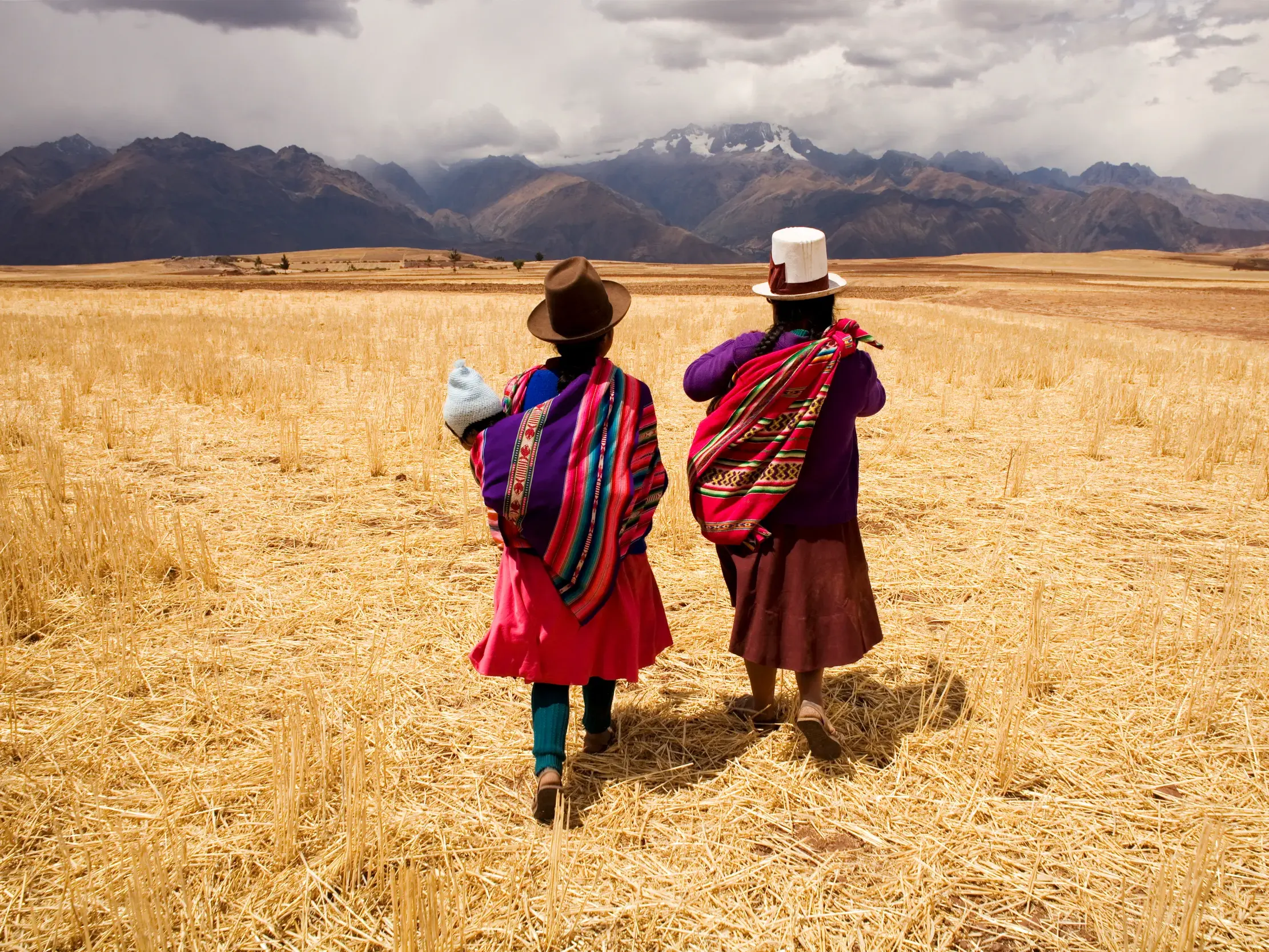 Zwei einheimische Frauen in bunter Tracht durchqueren ein Feld im Heiligen Tal vor der majestätischen Bergkette der Anden.