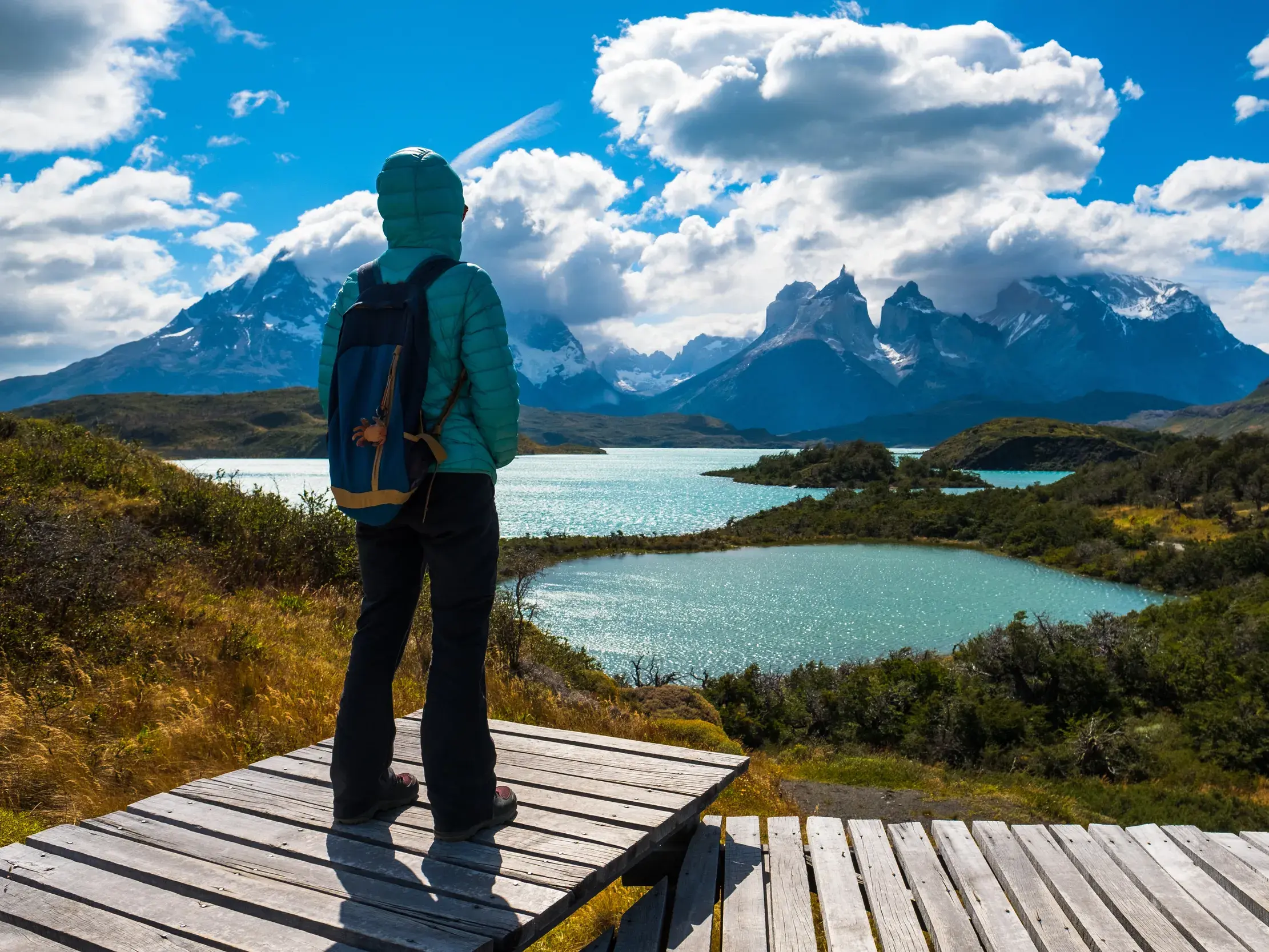 Wanderer auf einem Holzsteg blickt über einen türkisen See auf die markanten Gipfel des Torres del Paine Nationalparks.