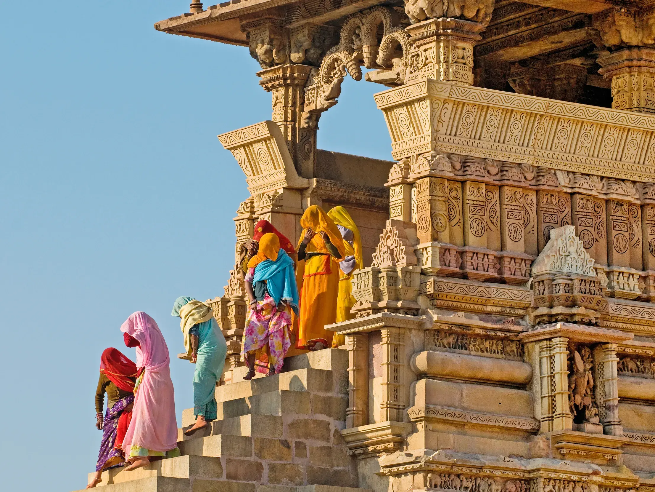 Gruppe indischer Frauen in farbenfrohen Saris auf der Steintreppe eines kunstvoll verzierten Sandsteintempels in Khajuraho.