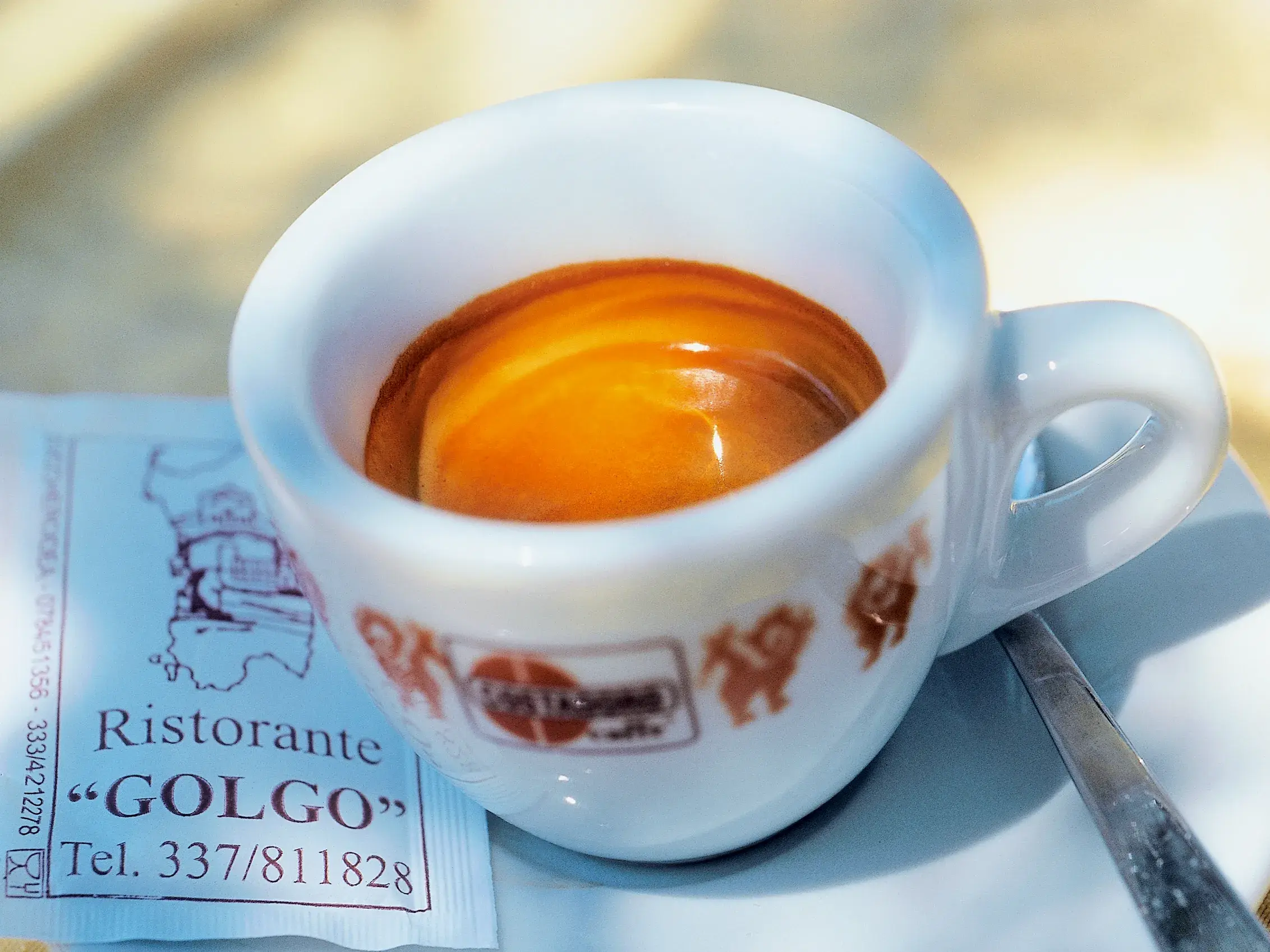 Nahaufnahme einer Tasse Espresso mit goldener Crema neben einem Zuckerpäckchen vom Ristorante Golgo auf Sardinien.