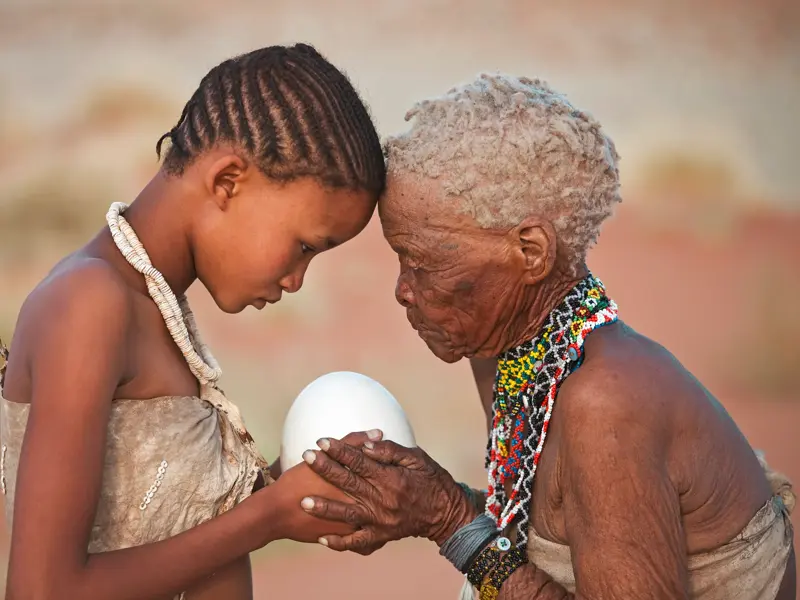 Eine ältere San-Frau und ein junges Mädchen berühren ihre Stirnen und halten gemeinsam ein Straußenei in der Kalahari-Wüste.
