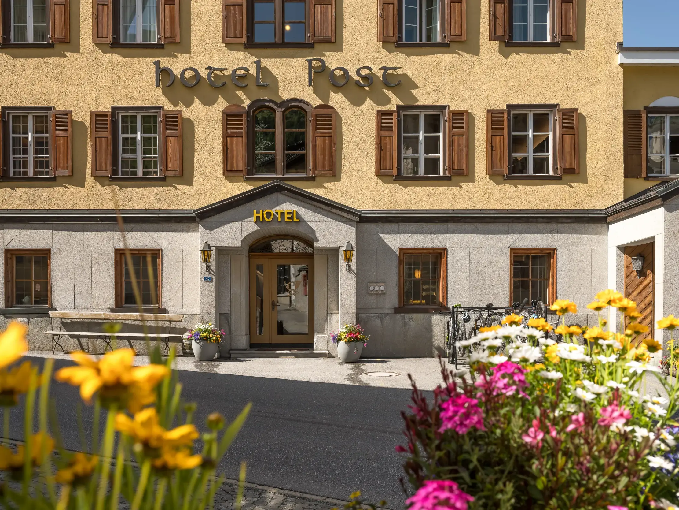 Die sonnige Fassade des Hotel Post im Bergdorf Bivio mit Holzfensterläden und bunten Sommerblumen im Vordergrund.