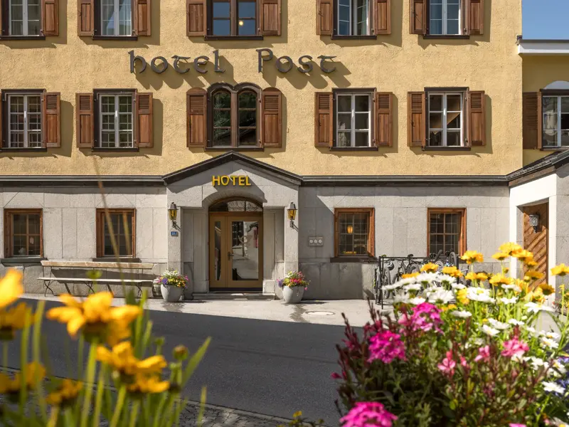 Die sonnige Fassade des Hotel Post im Bergdorf Bivio mit Holzfensterläden und bunten Sommerblumen im Vordergrund.