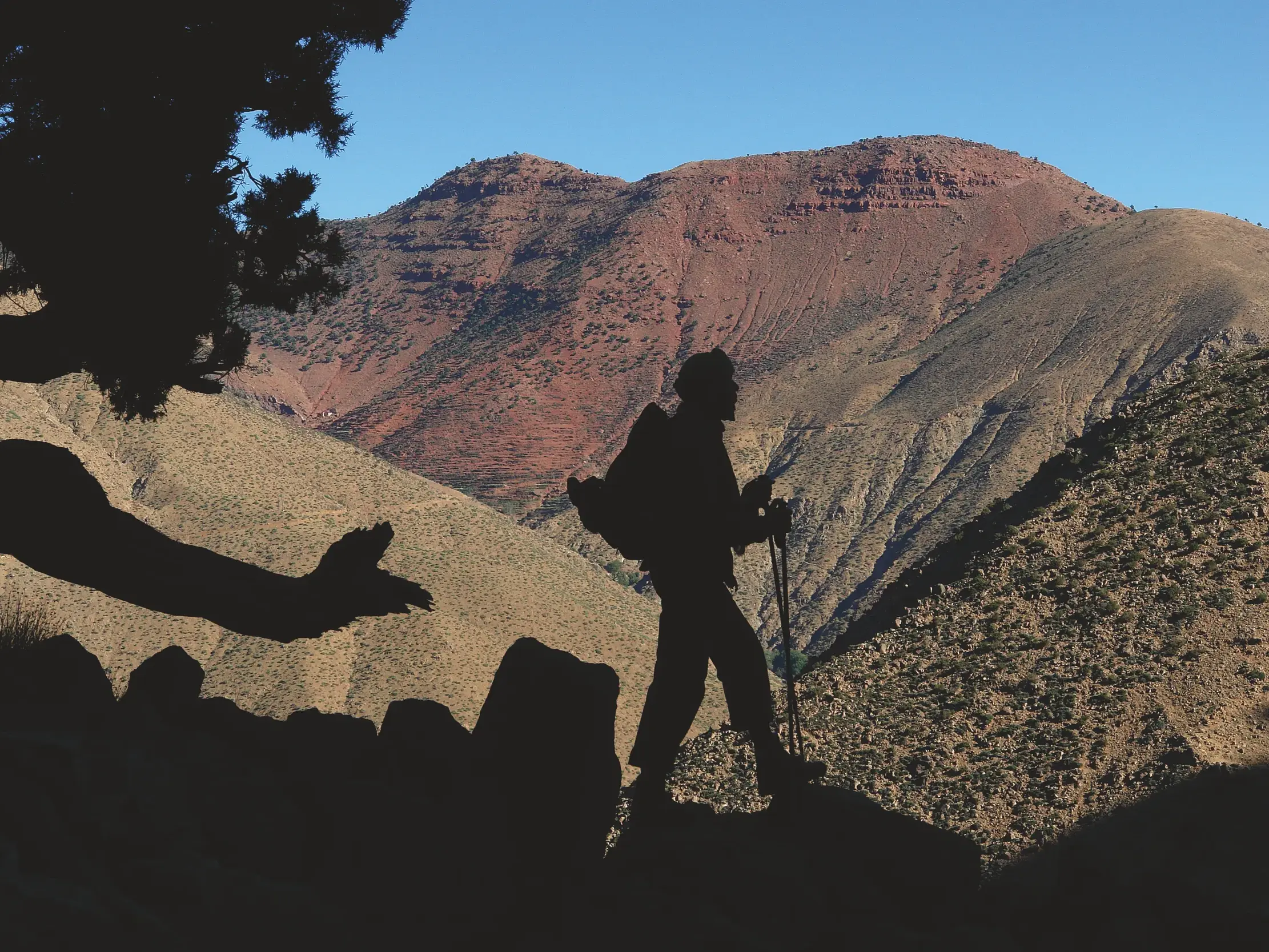 Trekking im Hohen Atlas: Silhouette eines Wanderers mit Stöcken vor der Kulisse der rötlichen Berglandschaft Marokkos.