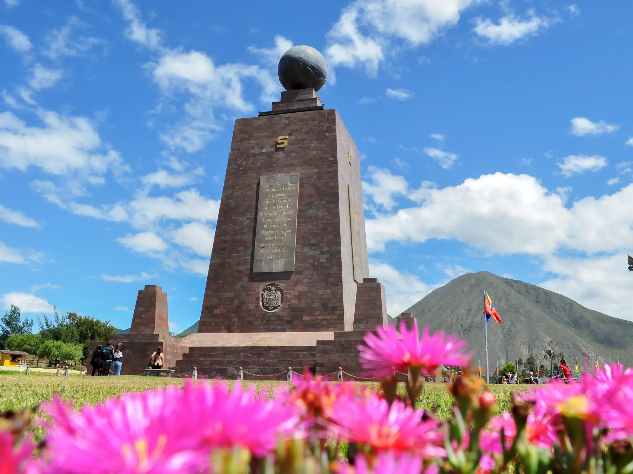 Das Äquatordenkmal Mitad del Mundo in Quito an einem sonnigen Tag, mit leuchtend pinken Blumen im Vordergrund und einem Berg im Hintergrund.
