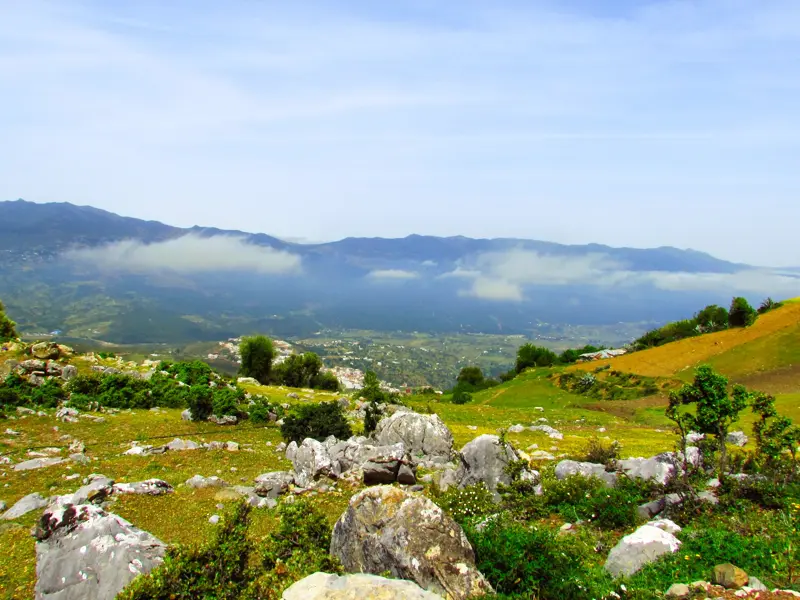 Panoramablick beim Wandern über ein grünes Tal auf die wolkenverhangene Bergkette des Rif-Gebirges in Nordmarokko.