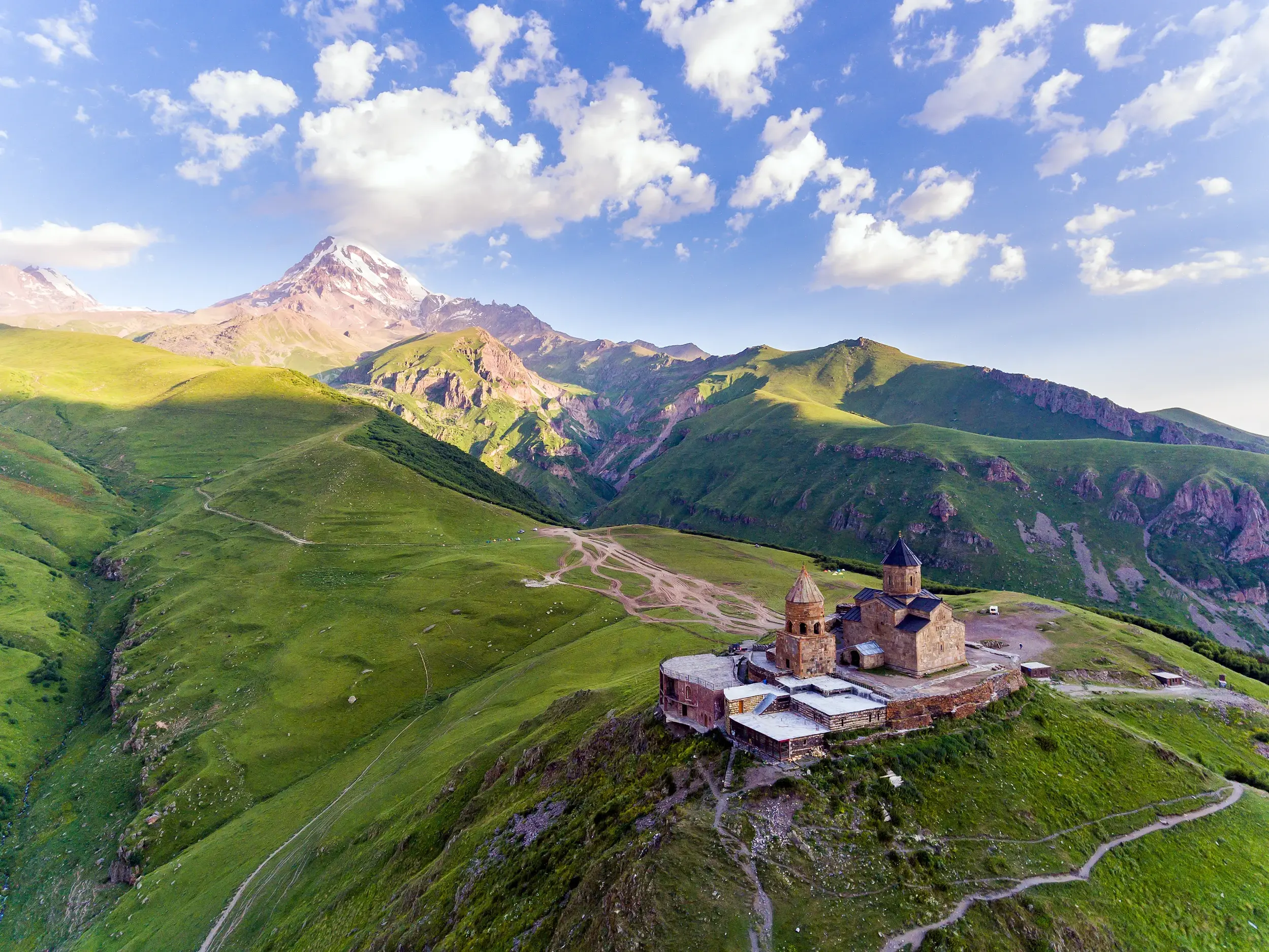 Die steinerne Gergetier Dreifaltigkeitskirche auf einem grünen Gipfel vor dem schneebedeckten Fünftausender Kasbek in Georgien.