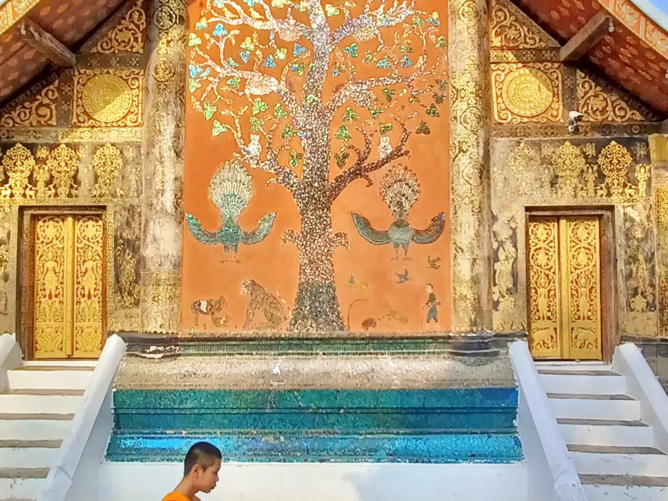 Prunkvolles Glasmosaik des Lebensbaums an der Tempelfassade des Wat Xieng Thong in Luang Prabang, flankiert von goldenen Toren.